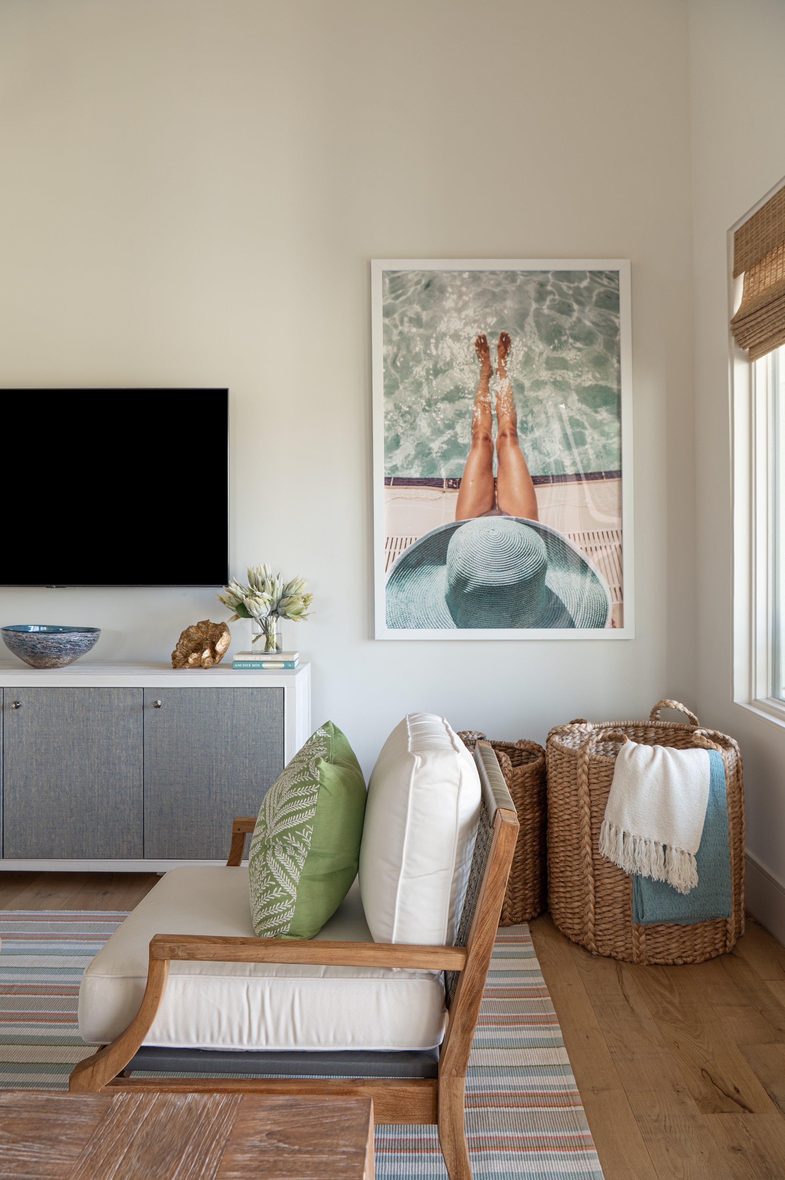 Living room with a white armchair with green and white pillows, wicker baskets, a wall art of a person relaxing by the water, a cabinet, TV, and various decorations.