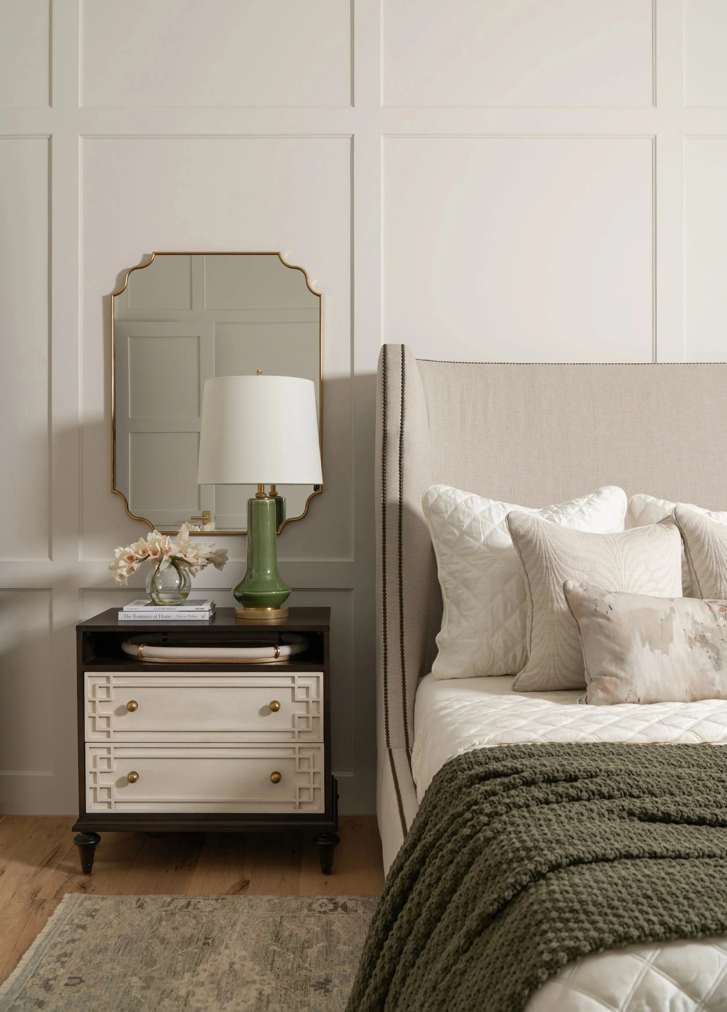 A bedroom scene with a beige upholstered bed, white pillows, a dark green blanket, a side table with a white lamp and a vase of flowers, a mirror, and a light-colored rug.