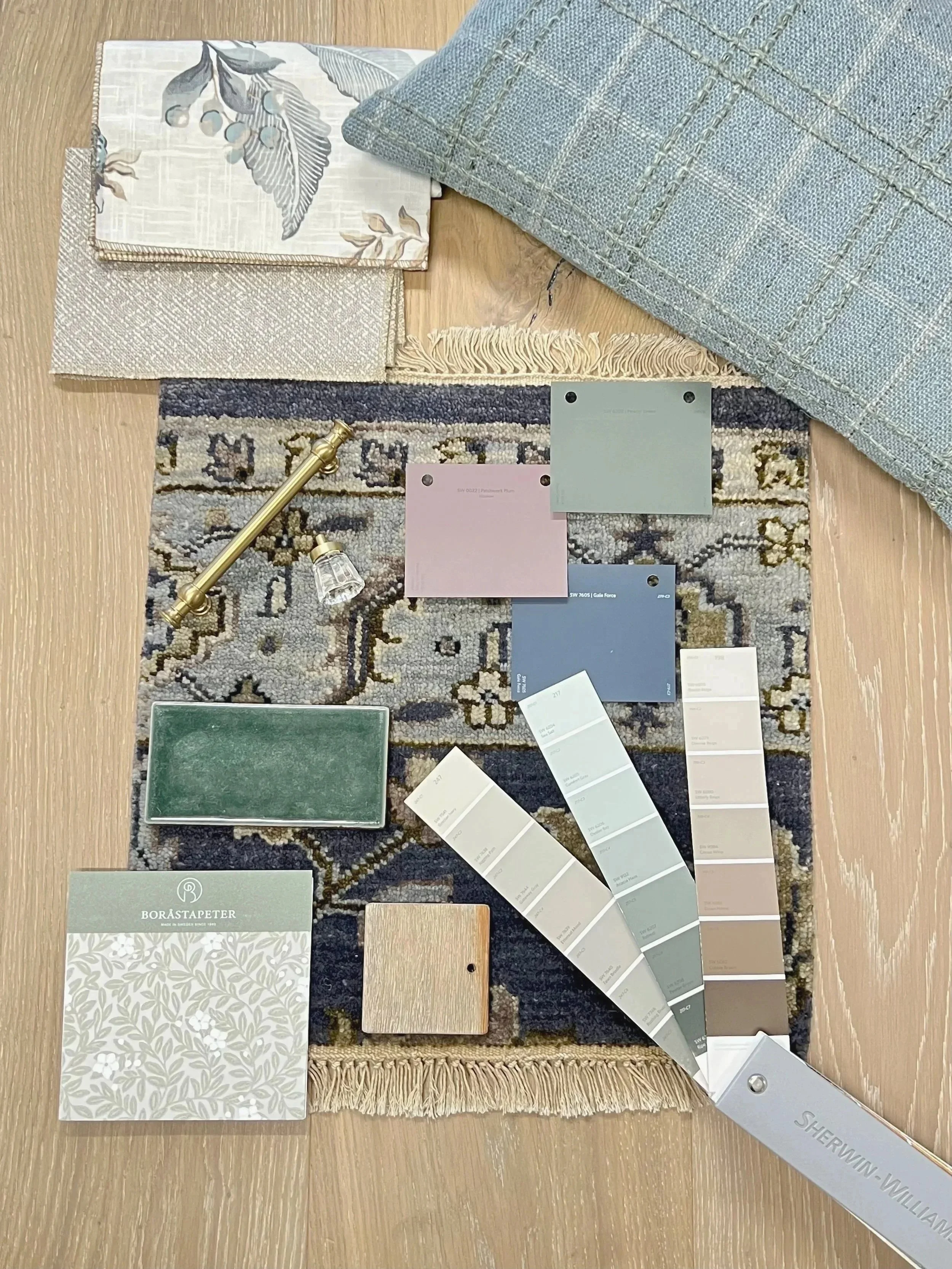 A variety of home decor and design materials arranged on a wooden surface, including fabric samples, paint color swatches, tile samples, and a small sample of wallpaper. The items are organized with some overlapping and a large metal sample puller labeled 'SHERWIN-WILLIAMS' holding some of the paint color swatches.