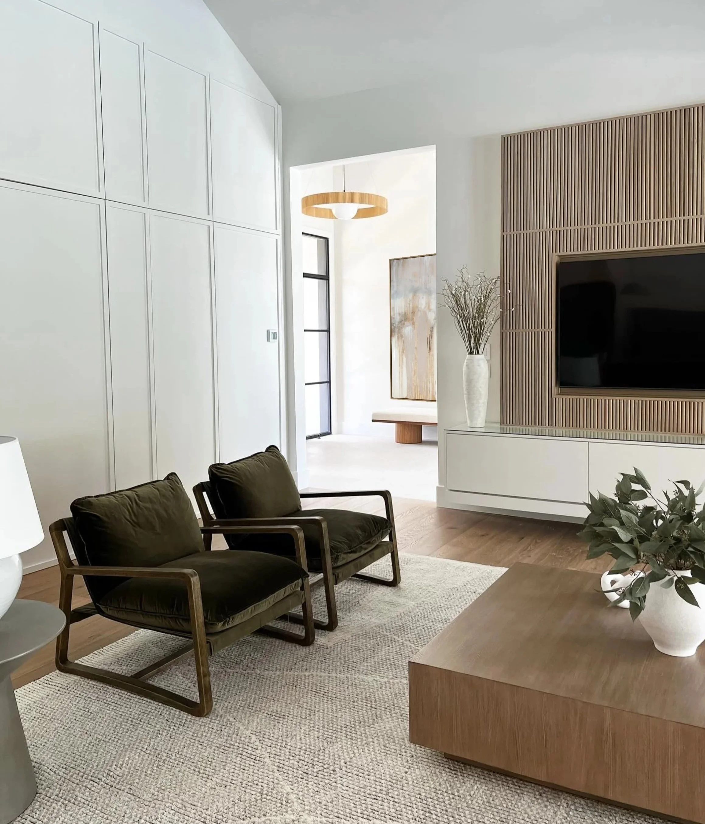 Living room with black armchairs, a light-colored rug, a wooden coffee table, a large wall-mounted television, and a white vase with branches. The room has white walls, a wooden slatted divider, and a modern ceiling light. Natural light enters through a window.