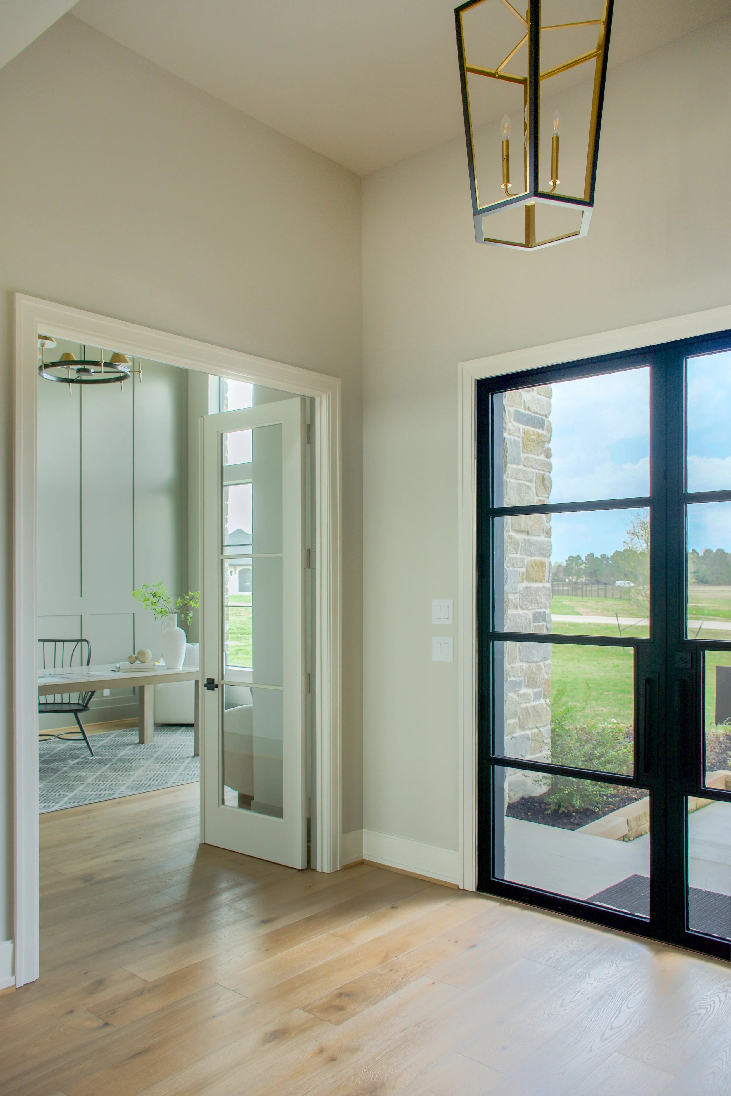 View of a home interior with a wooden floor, white walls, a black framed glass door leading outside, and a modern ceiling light fixture. Through an open door, a dining area with a table, chairs, and a decorative vase is visible.