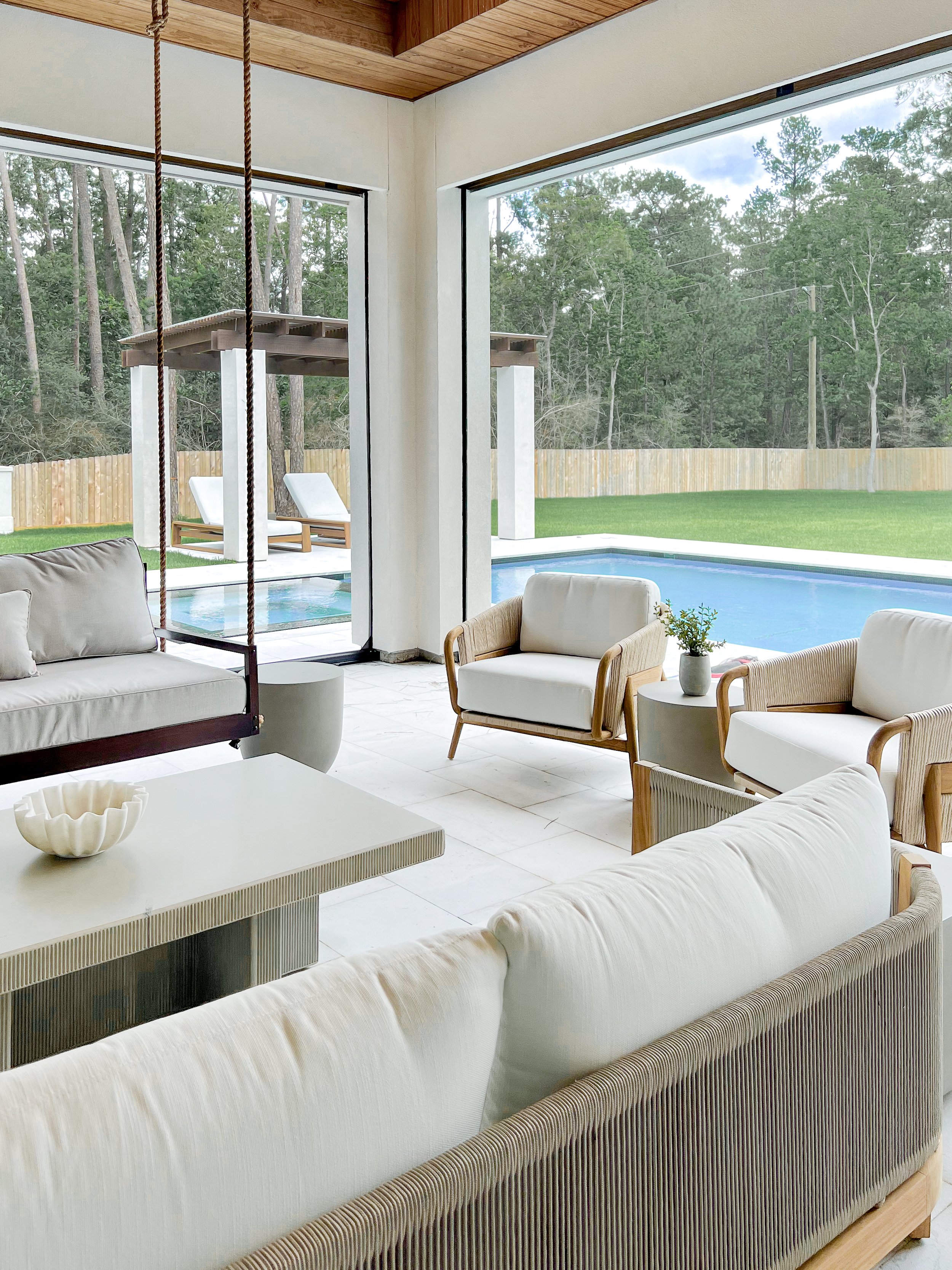 Living room with white cushioned furniture, a coffee table with a decorative bowl, large glass doors revealing a backyard with a grass lawn, pool, lounge chairs under a wooden pergola, and trees in the background.