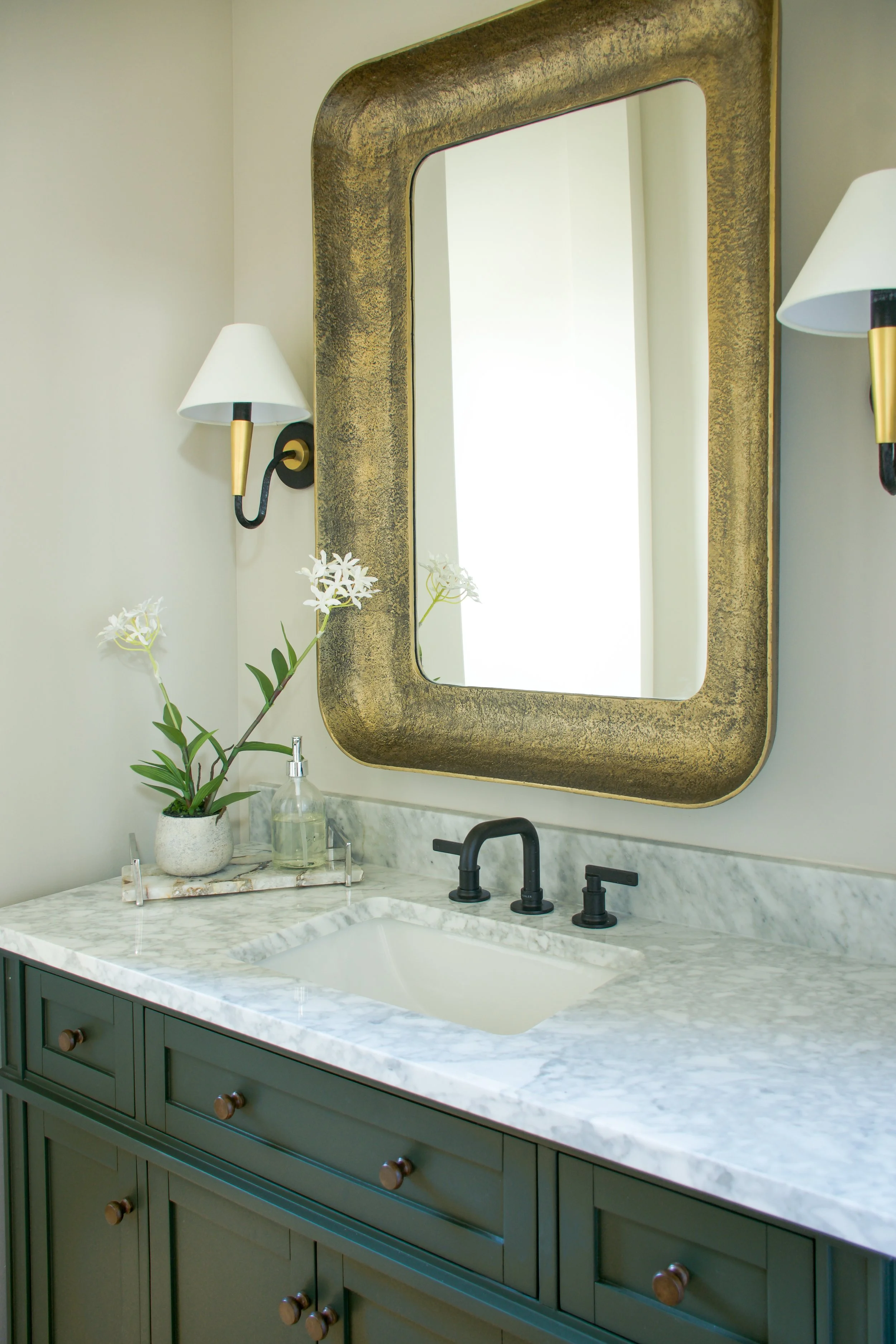 A bathroom vanity with dark green cabinetry, a marble countertop, and a white undermount sink with black faucet fixtures. Above, a large gold-framed mirror is mounted on the wall. To the left, there are two wall-mounted lamps with white shades and go