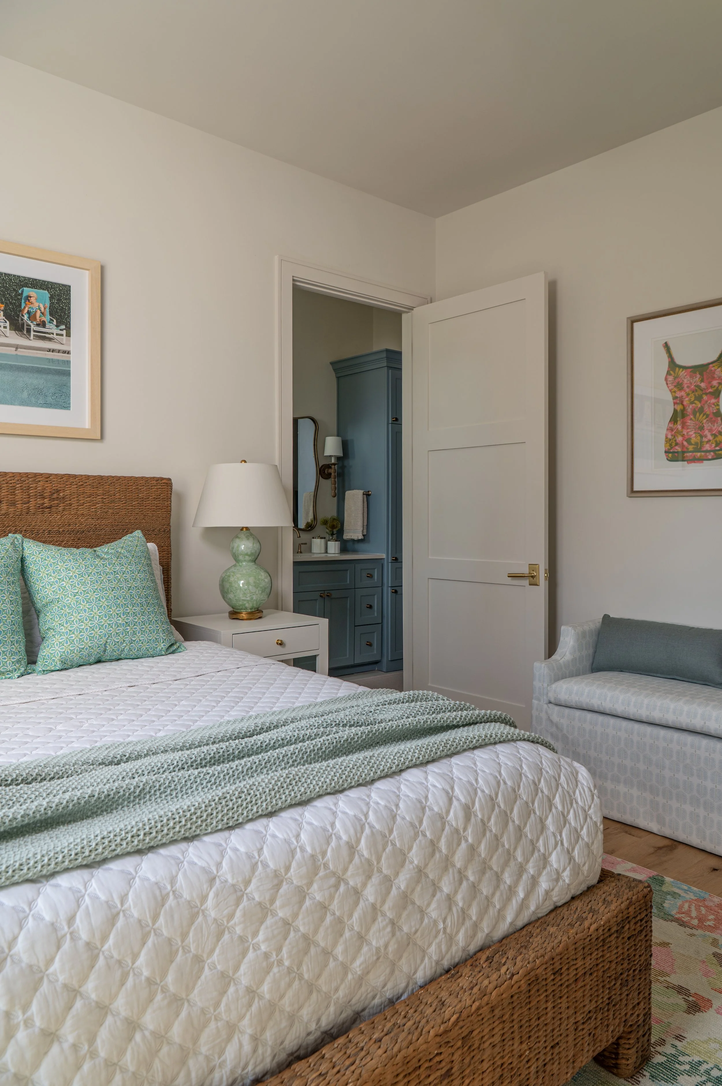 A bedroom with a bed, pillows, a bedside table with a green ceramic lamp, artwork on the wall, and a small blue vanity with a mirror in the background.