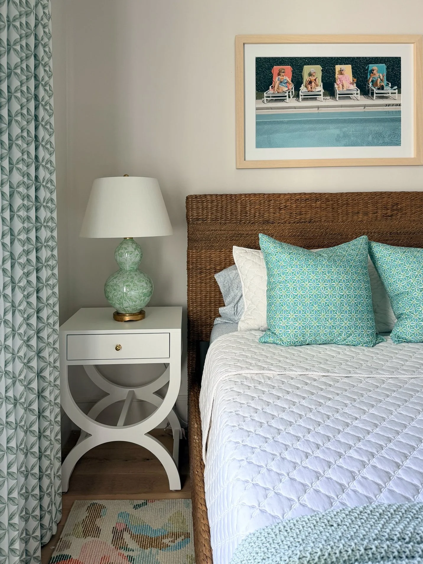 A bedroom corner with a bed having a wicker headboard, decorated with blue patterned pillows and a white quilt. Next to the bed is a white side table with a decorative green and white ceramic lamp. On the wall above, there's a framed painting of four