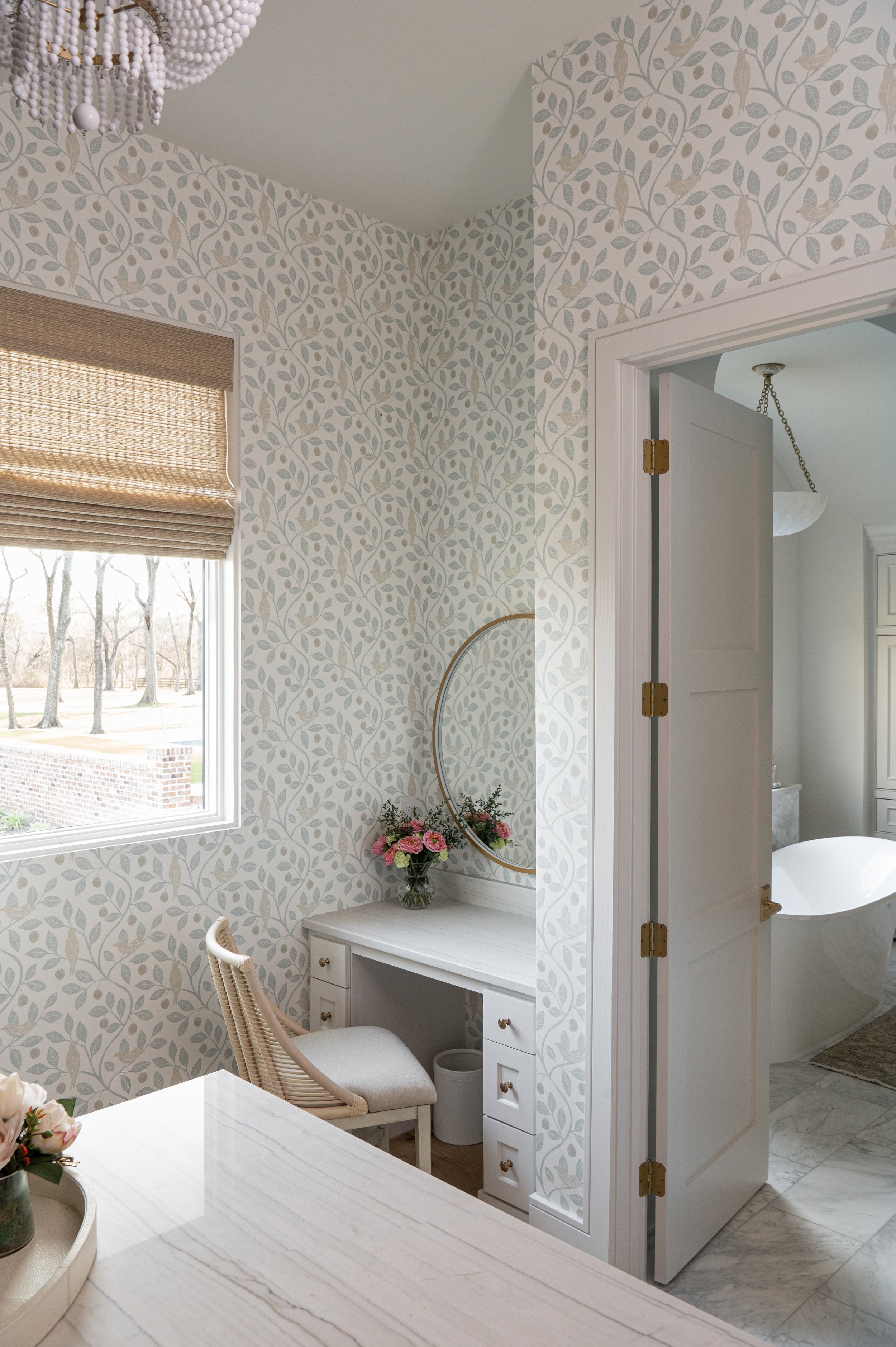 A corner of a room with wallpaper featuring a leafy pattern and bird designs, a window with a woven shade, a small white desk with a flower arrangement, a wicker chair, and a view into a bathroom with a freestanding bathtub visible through an open do