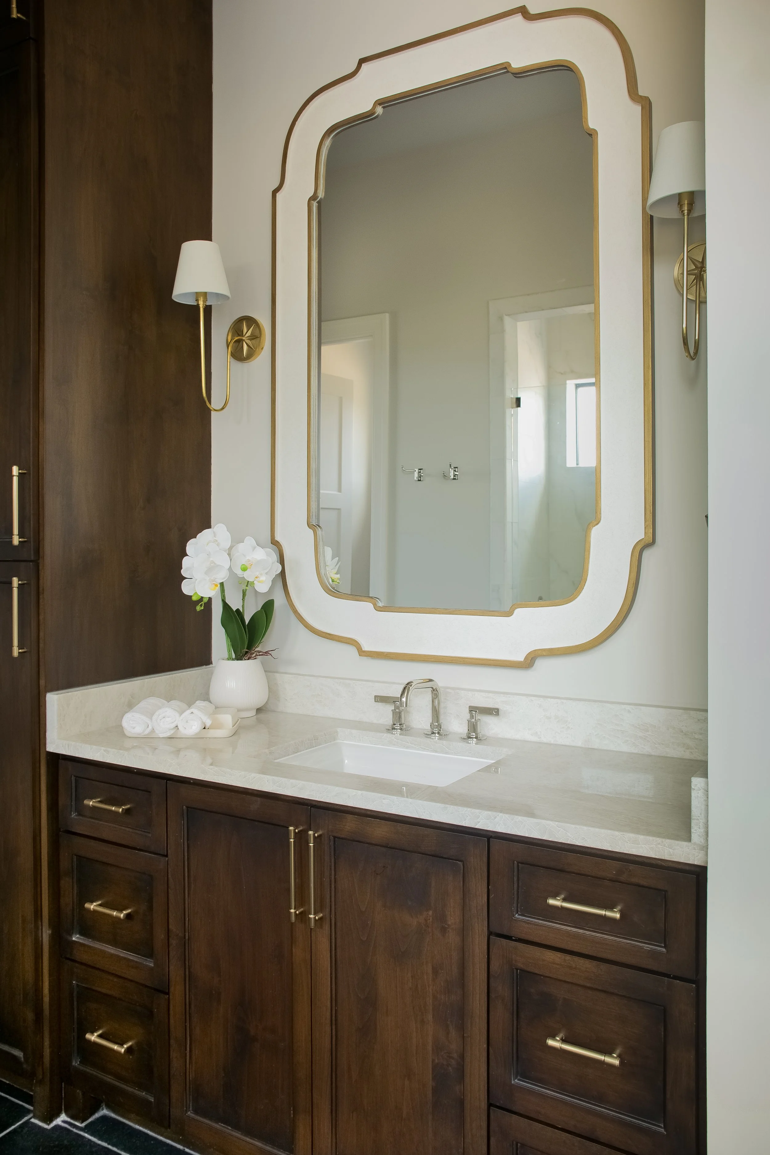 Bathroom vanity with dark wood cabinets, a white countertop, and a built-in sink. A large mirror with a decorative gold frame is mounted above the sink, flanked by two wall-mounted white lamps with gold accents. On the countertop is a white vase with