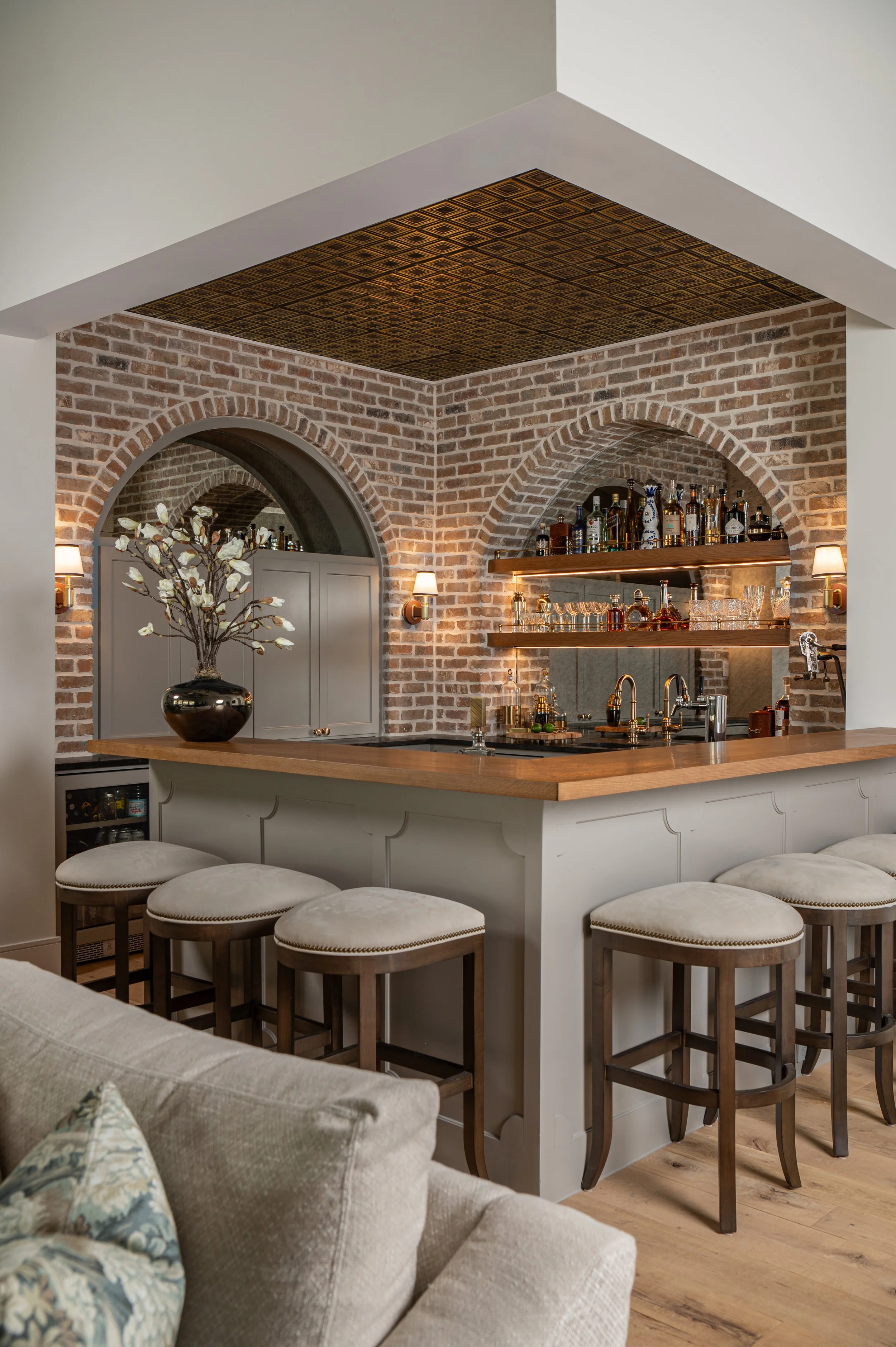 A kitchen with exposed brick walls, a bar counter with white upholstered stools, a vase with white flowers, and a ceiling feature with a pattern. There are bottles and glassware on open shelves behind the bar.