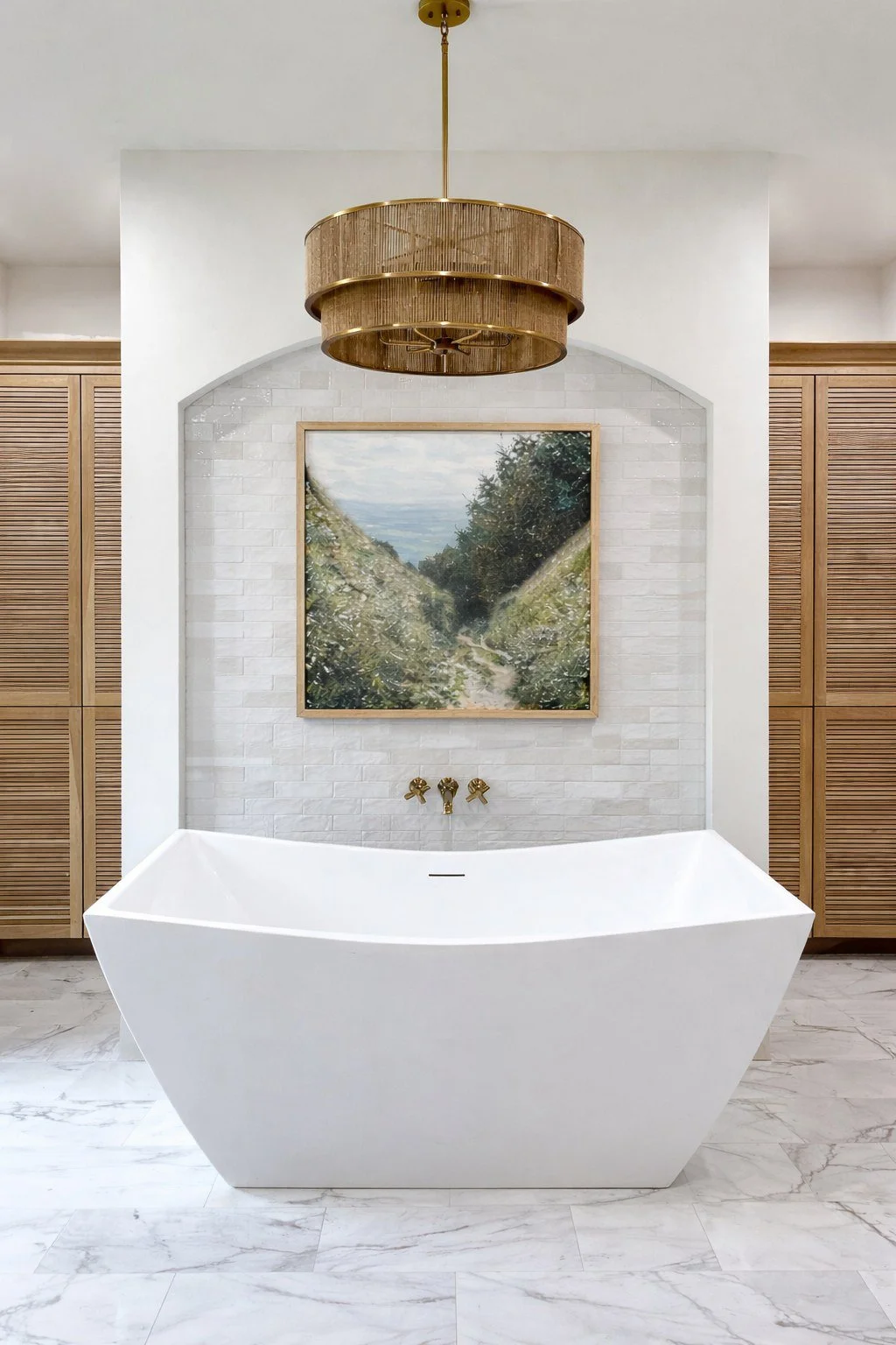 Modern bathroom with a white freestanding bathtub, a large landscape painting on a white tiled wall, gold fixtures, and a brass chandelier.
