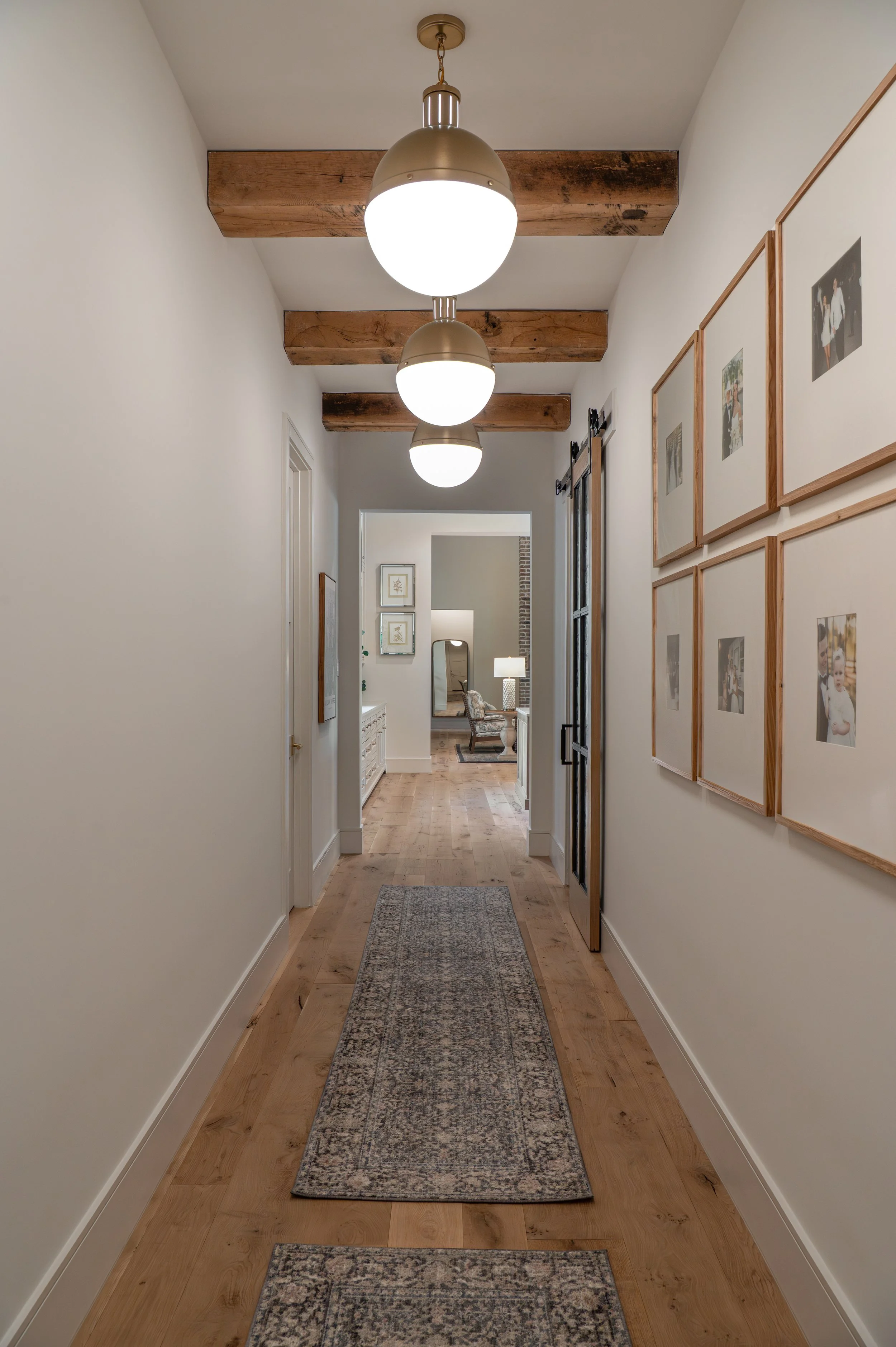 A hallway with white walls, three hanging globe lights, exposed wooden ceiling beams, framed photographs on the right wall, and hardwood flooring with a runner rug.
