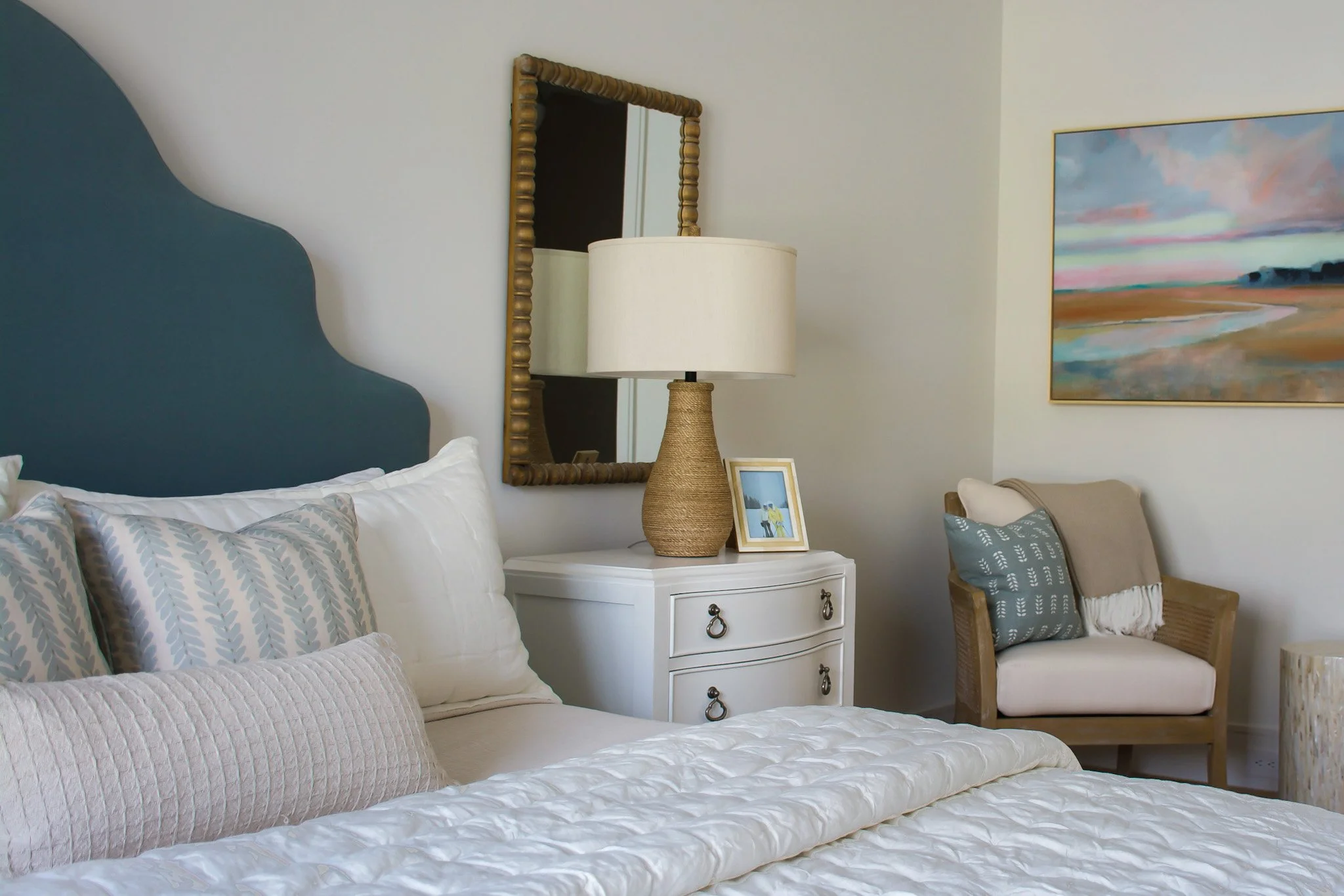 A bedroom with a white bed featuring decorative pillows, a white nightstand with a framed photo, a table lamp, a wicker chair with a pillow, a tall mirror with a wooden frame, and a colorful landscape painting on the wall.