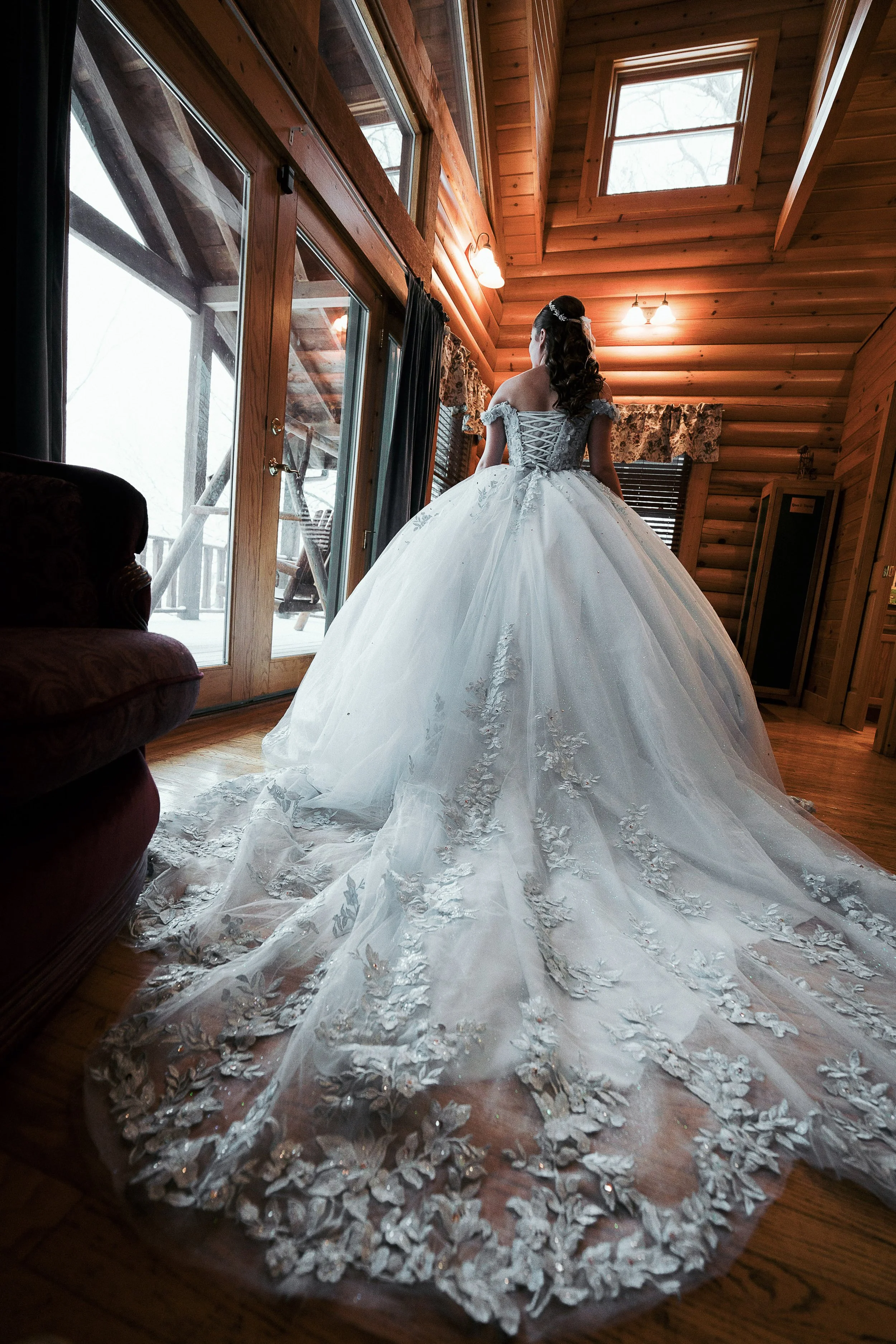 A bride in a white wedding gown standing inside a wooden cabin near large glass doors leading outside to a snowy landscape.