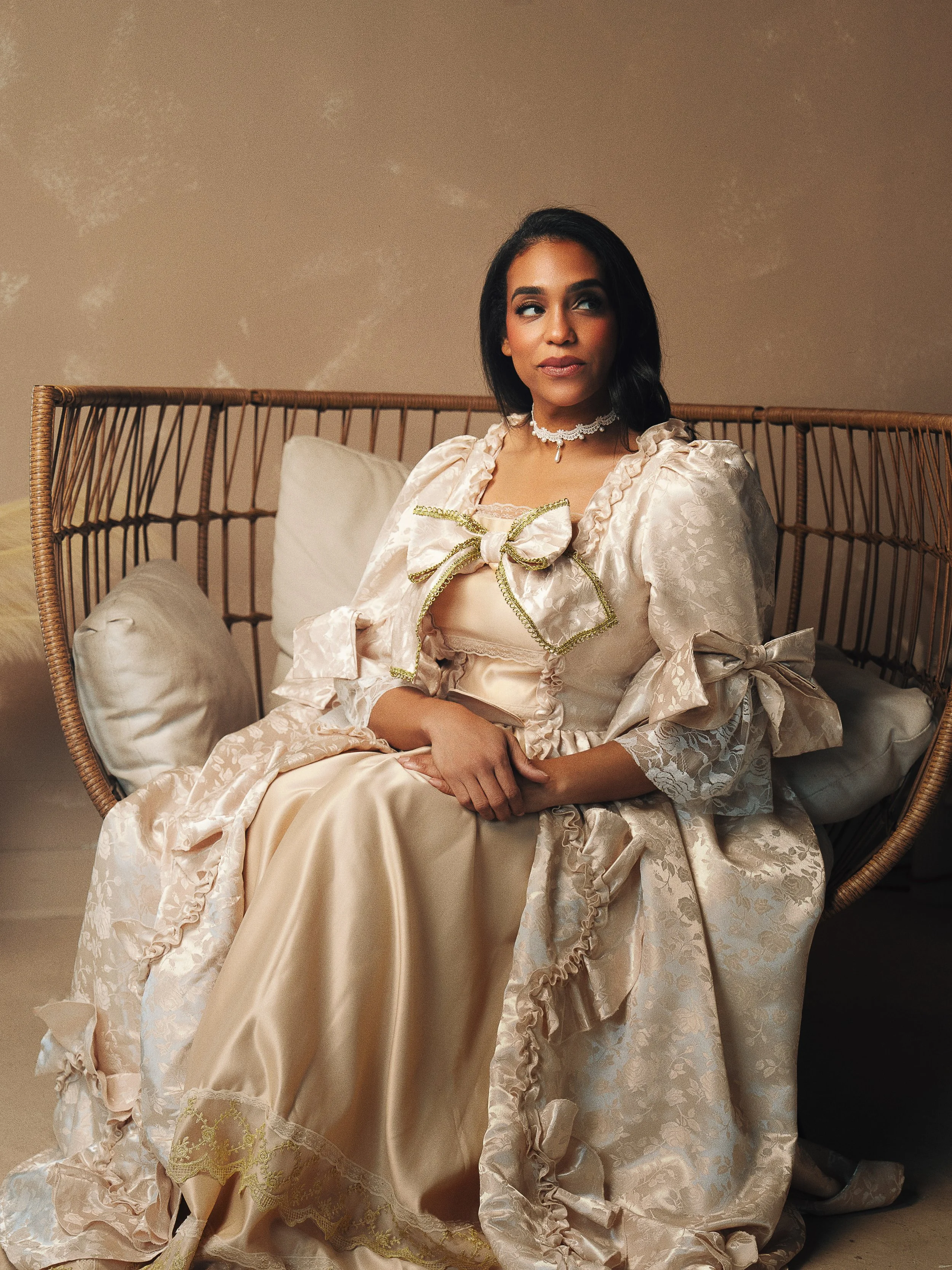 A woman dressed in an elegant, vintage-style cream-colored gown with lace and ribbon details, sitting on a wicker chair with beige pillows against a neutral background.