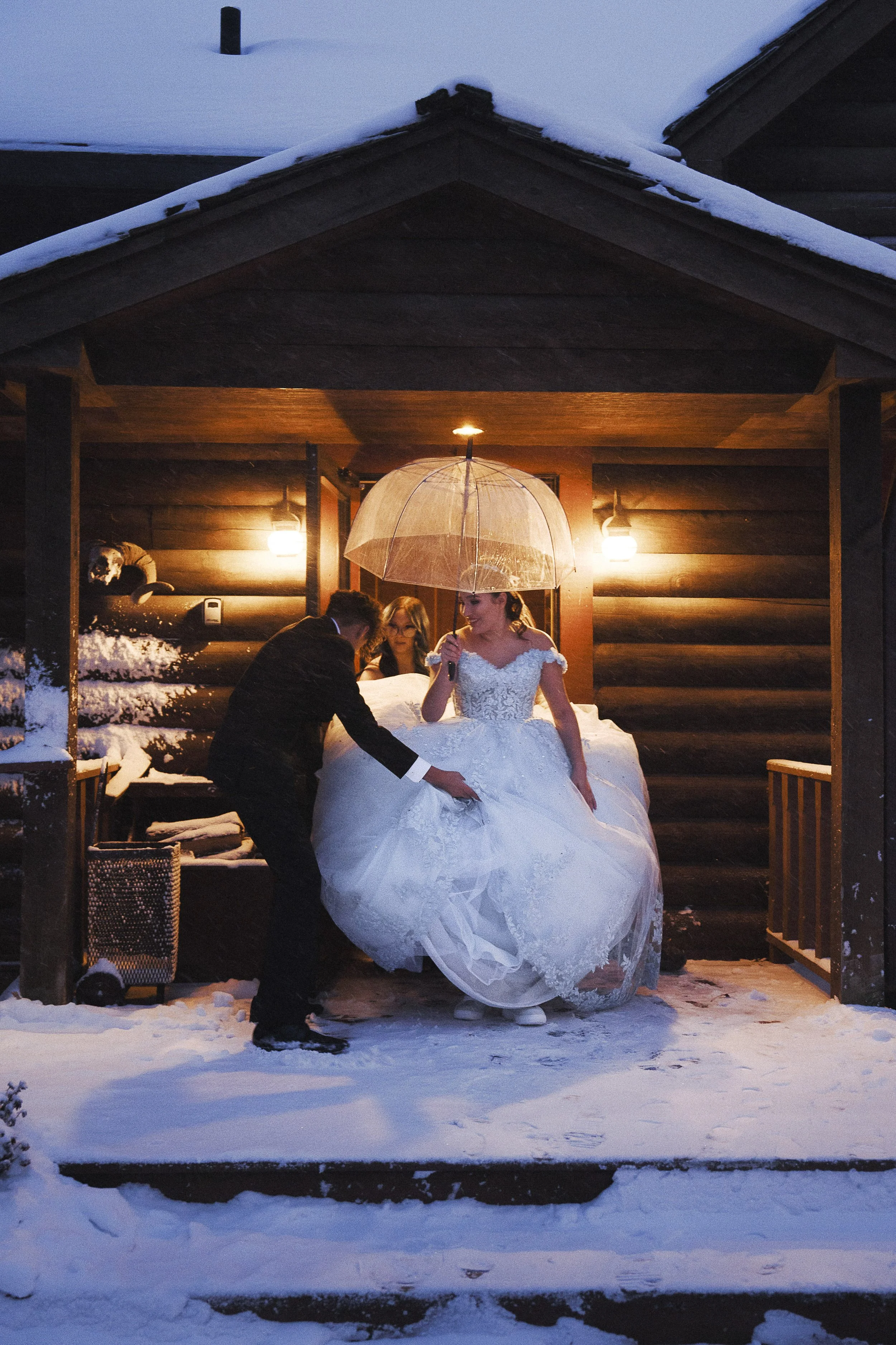 Bride under an umbrella, groom helping her with her dress outside a snow-covered cabin at night.