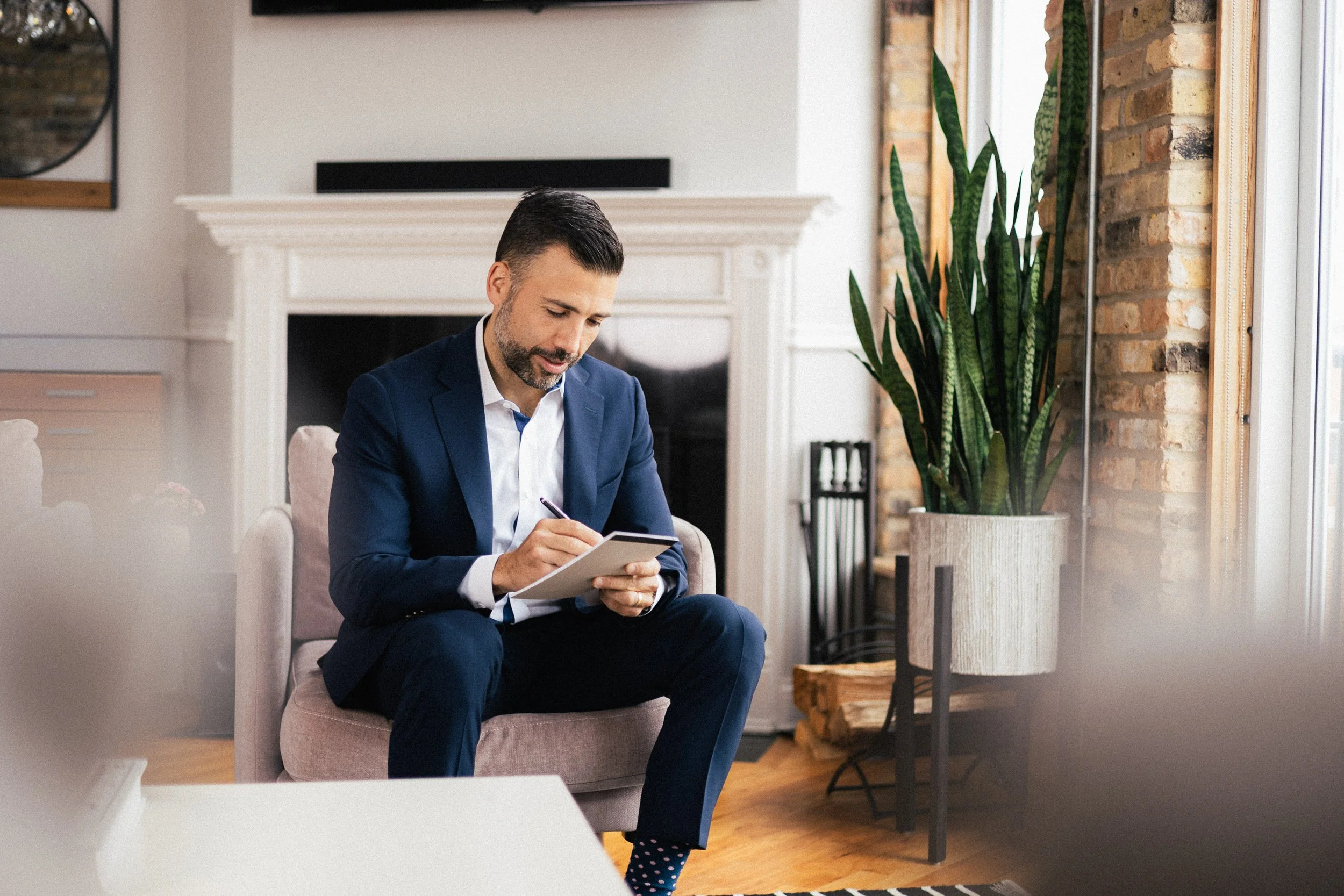 A groom in a blue suit sitting in a pink armchair in a modern living room, writing in a notebook with a pen. There is a large green plant next to him and a fireplace behind him.