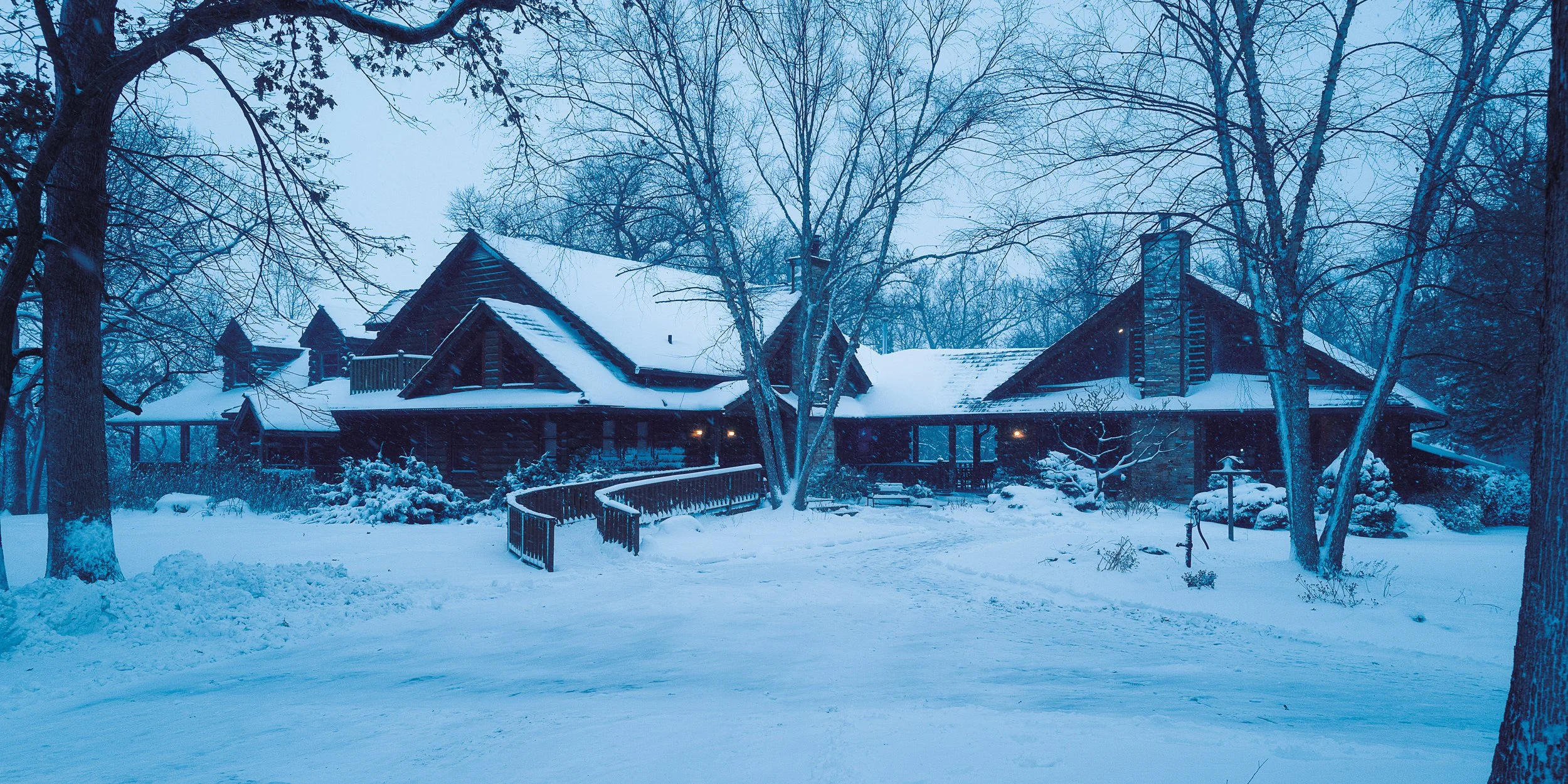 A wooden cabin surrounded by snow and bare trees during winter, with a small pathway and a wooden railing in the snow.