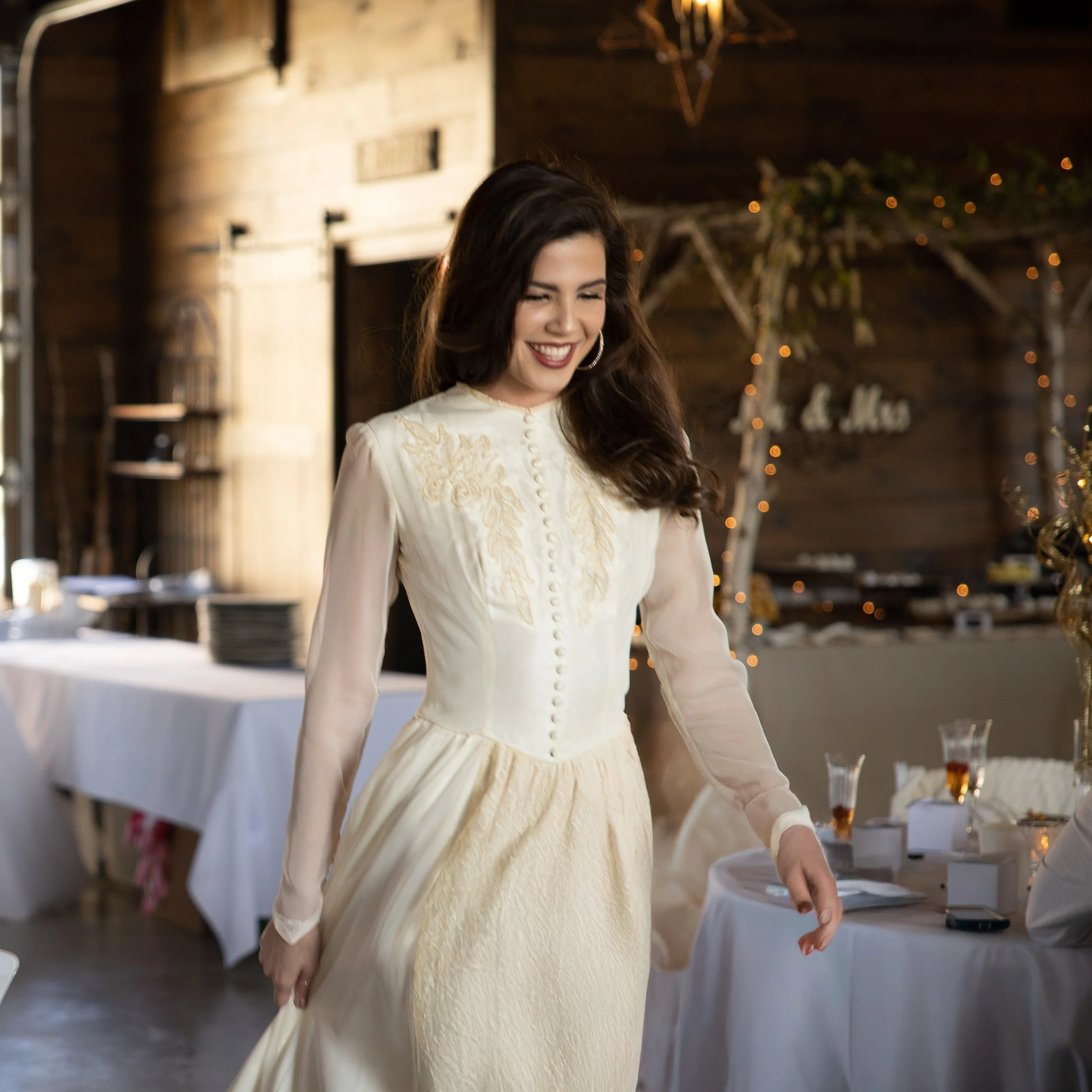 A woman smiling and wearing a vintage-style cream-colored wedding dress with embroidery details, standing in a rustic indoor setting decorated with fairy lights.