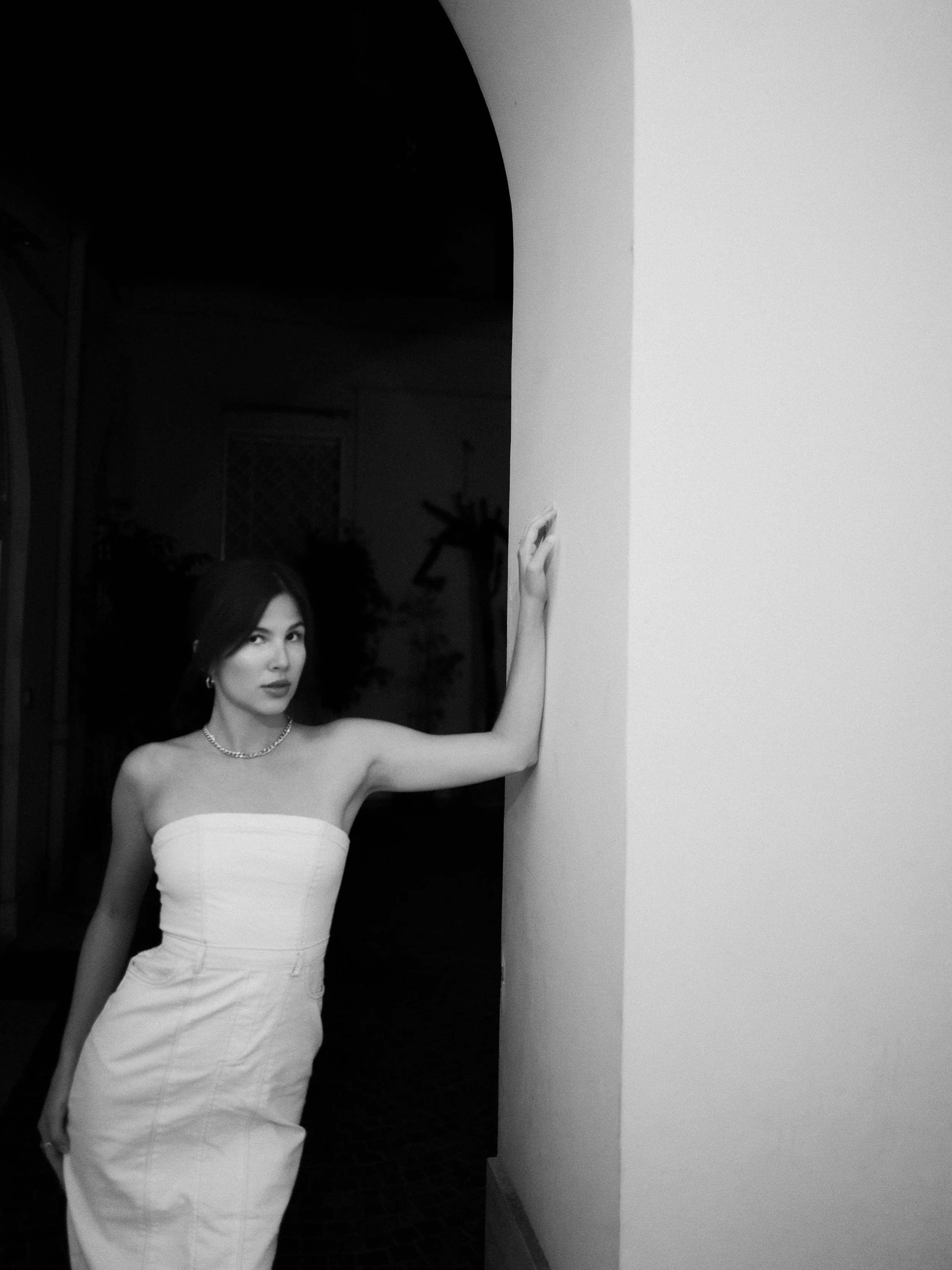 A bride in a strapless white weding dress with a necklace, standing against a European arch, with her arm extended and resting on the wall.