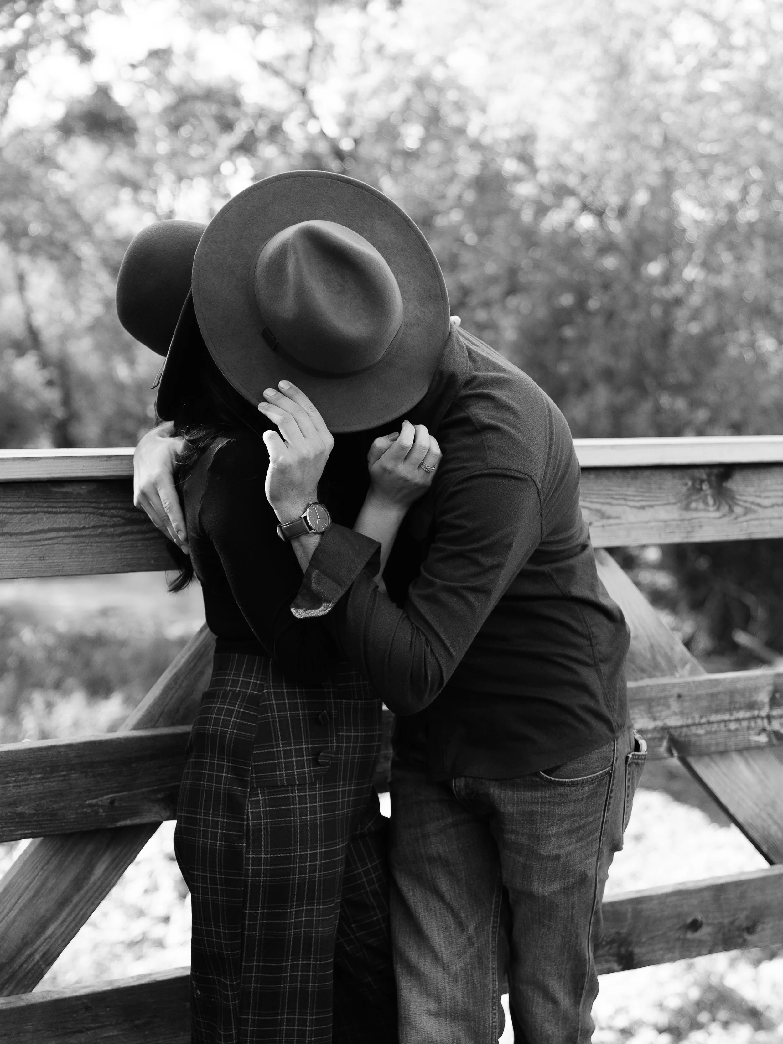 A black and white photo of a couple kissing behind a large hat, standing outdoors near a wooden fence and trees.