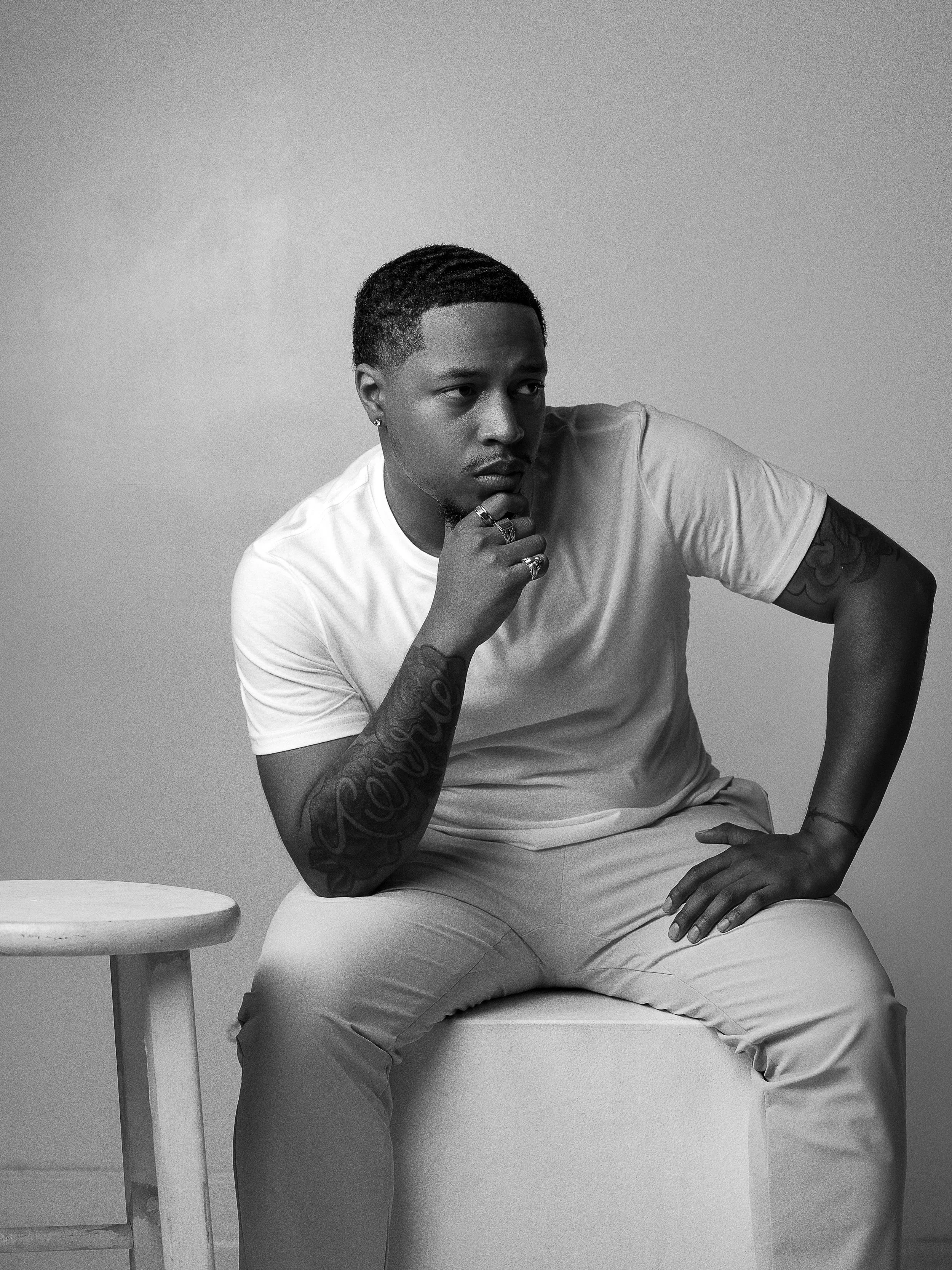 A black-and-white photo of a young man sitting on a stool with a contemplative expression, wearing a light-colored t-shirt and light pants. He has tattoos on both arms and rings on his fingers, with his right hand resting on his thigh and his left hand touching his chin.