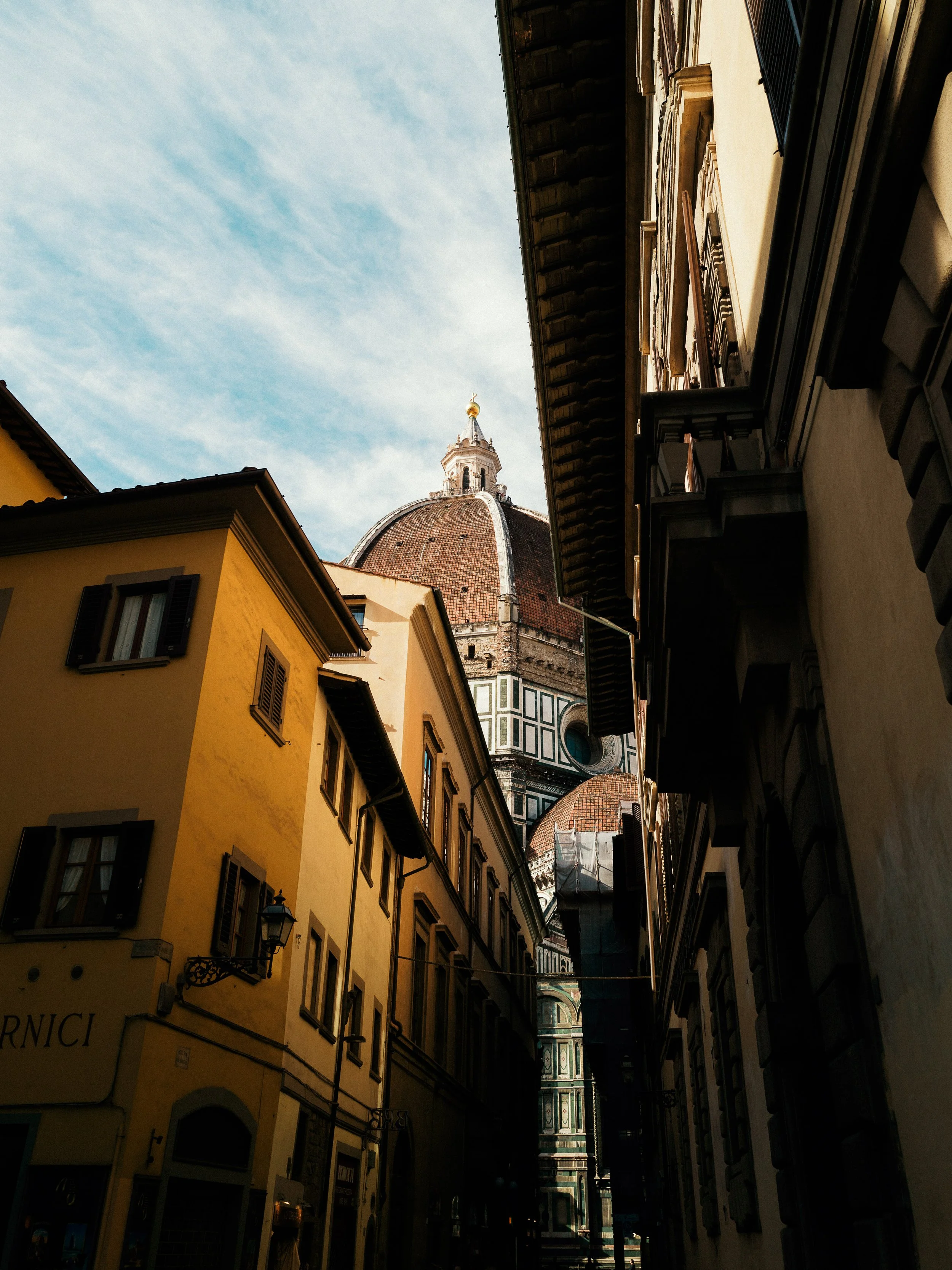 View of a narrow street between yellow and beige buildings with the Florence Cathedral's dome in the background under a partly cloudy sky.