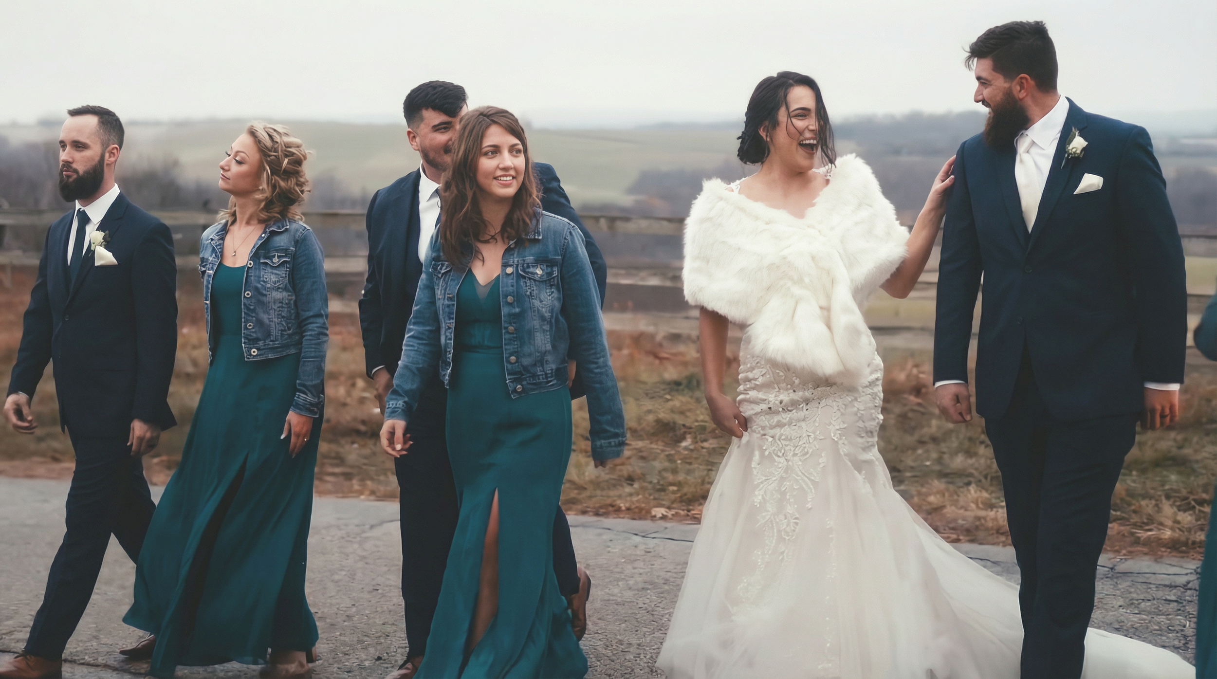 A bride in a white wedding gown and fur shawl walking arm-in-arm with a groom in a dark suit and white shirt, smiling and looking at each other, surrounded by the wedding party outdoors on a cloudy day.