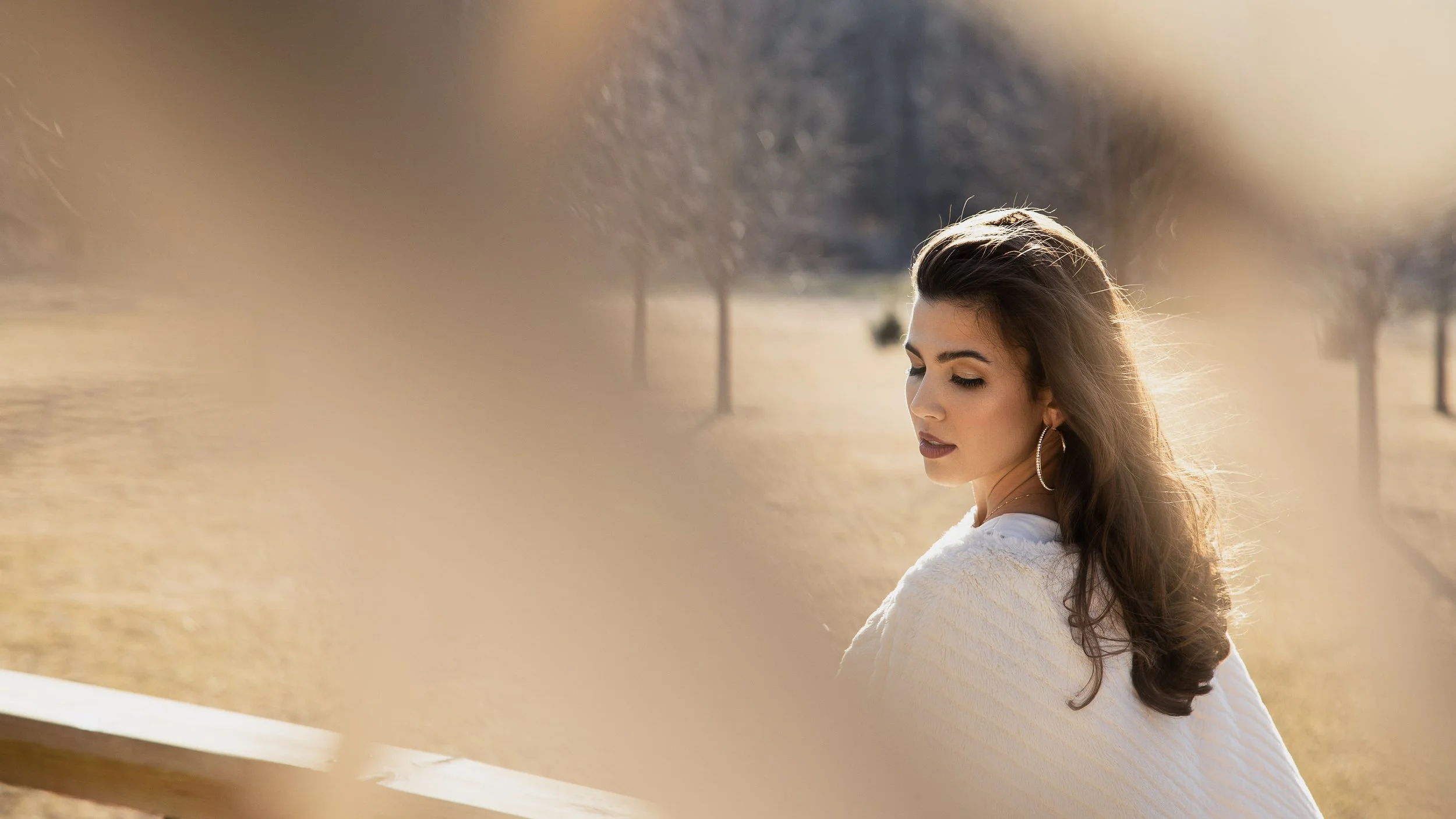 A bride with long brown hair and hoop earrings stands outdoors, closed eyes, wearing a white textured sweater, in a park with trees and a pathway in the background during sunset.