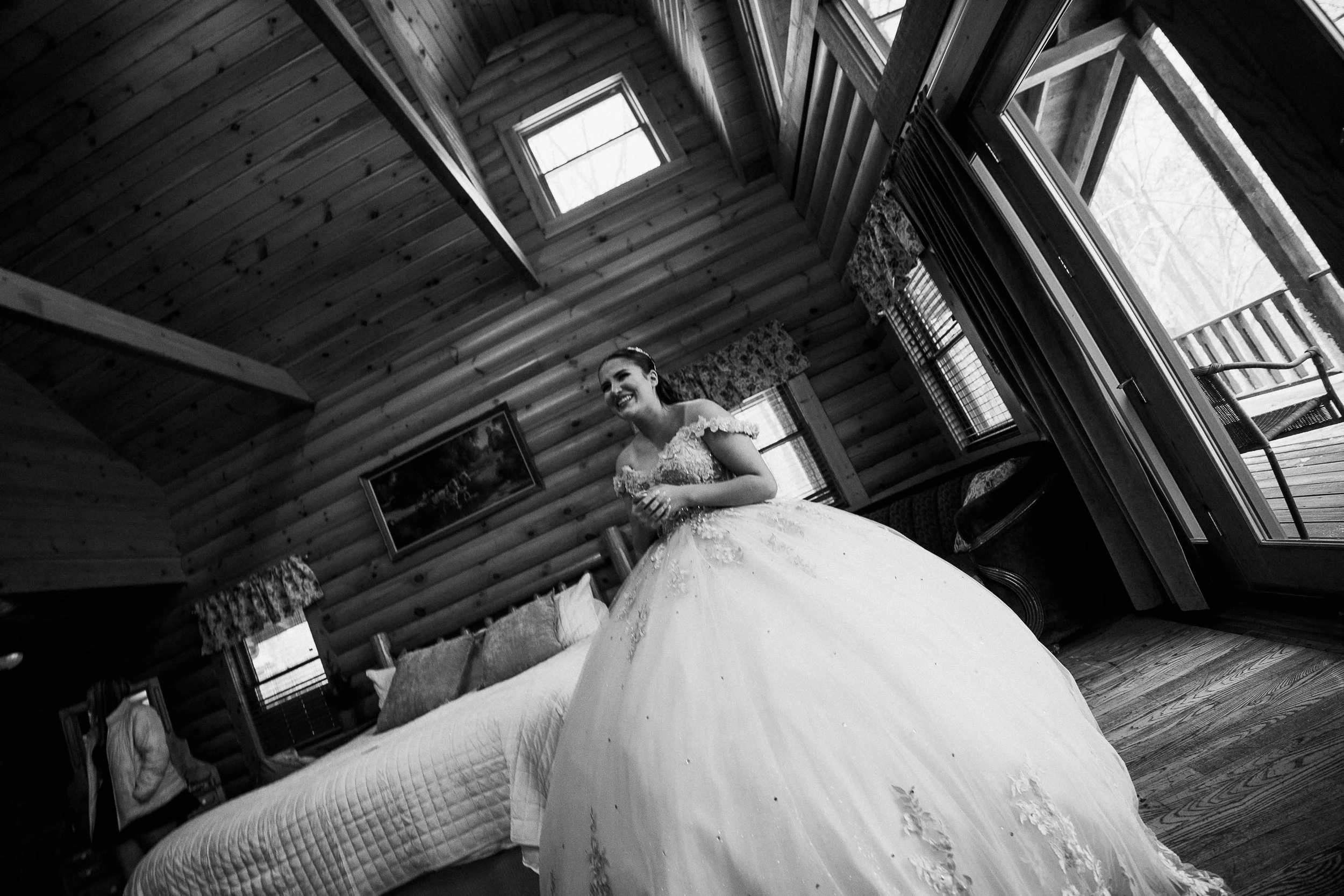 A bride in a wedding gown smiling inside a log cabin with wooden walls, windows, and furniture.