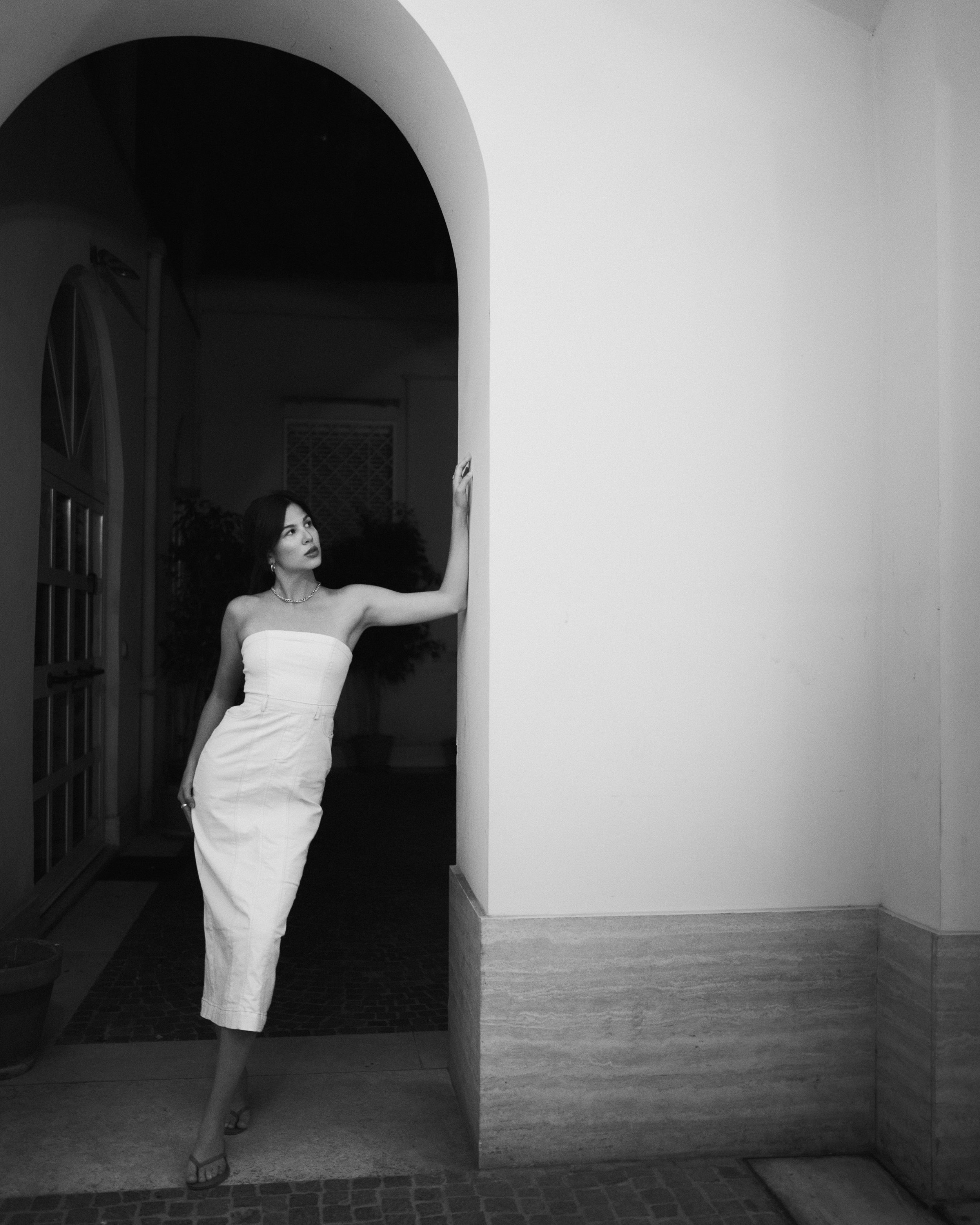 A bride in a strapless white wedding dress and high heels poses with her right hand on a wall in a European archway, looking to her left.