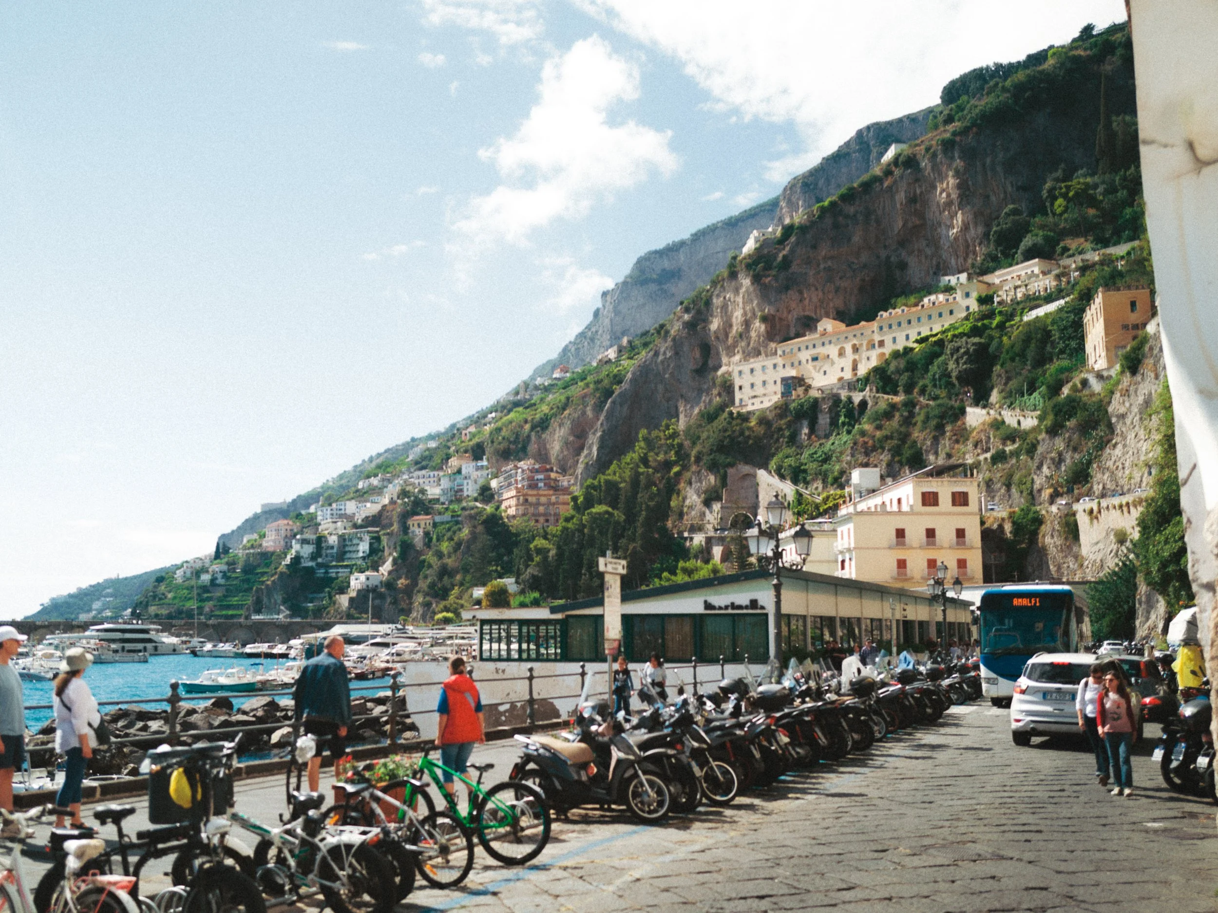 People walking along a harbor with parked motorcycles and bicycles, a bus, and a hillside with colorful buildings and lush greenery in the background. This is an elopement