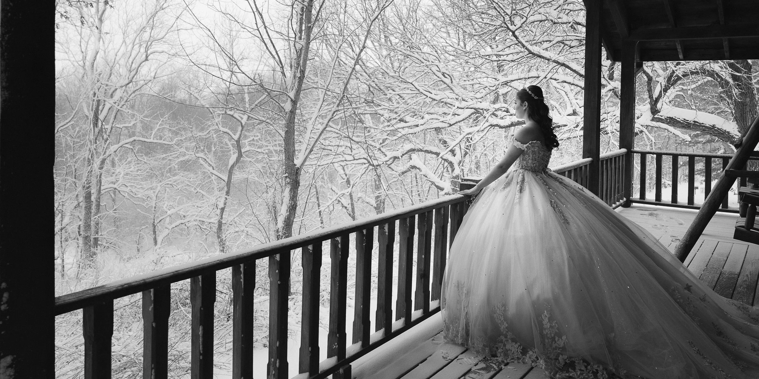 A bride in an ornate wedding dress stands on a snow-covered wooden porch, overlooking a snowy forest.