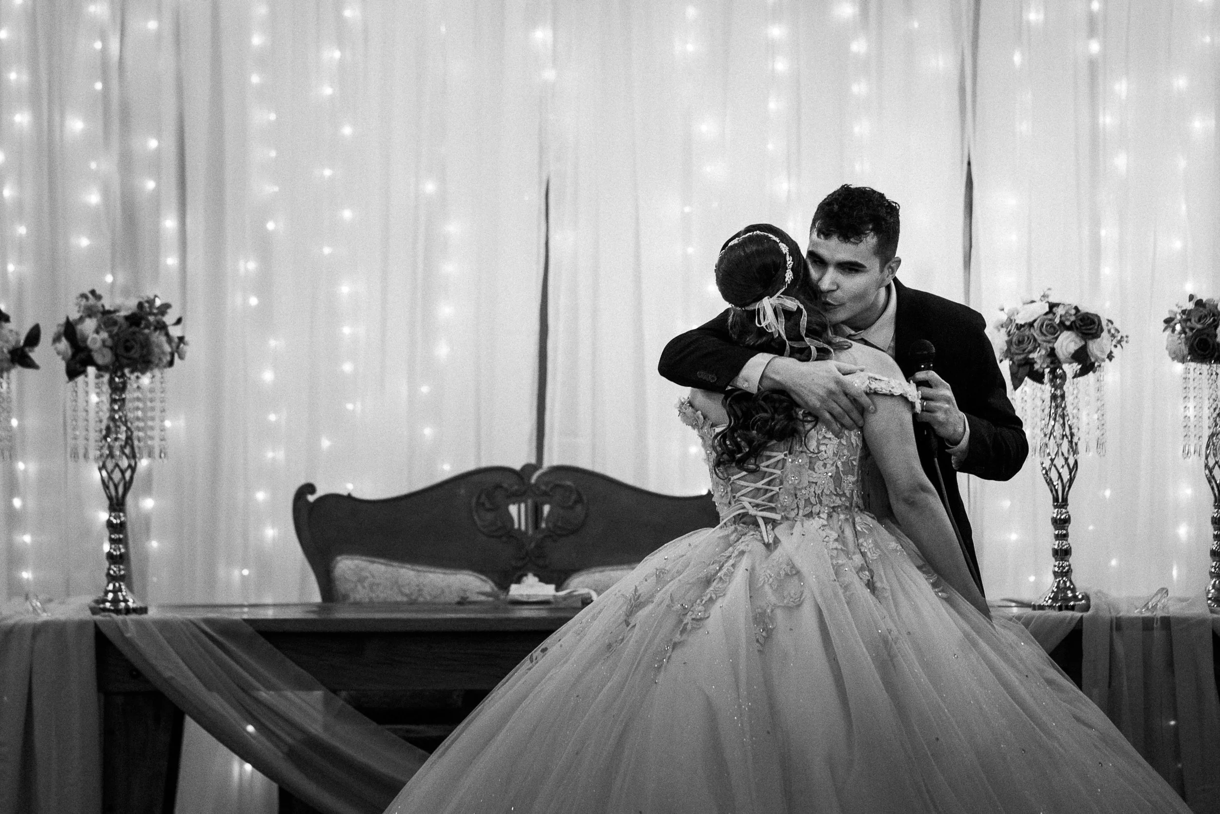 A wedding scene with a bride in a lace and embroidered gown and a groom in a dark suit sharing an embrace. The background features draped curtains with string lights and floral arrangements on tall stands.