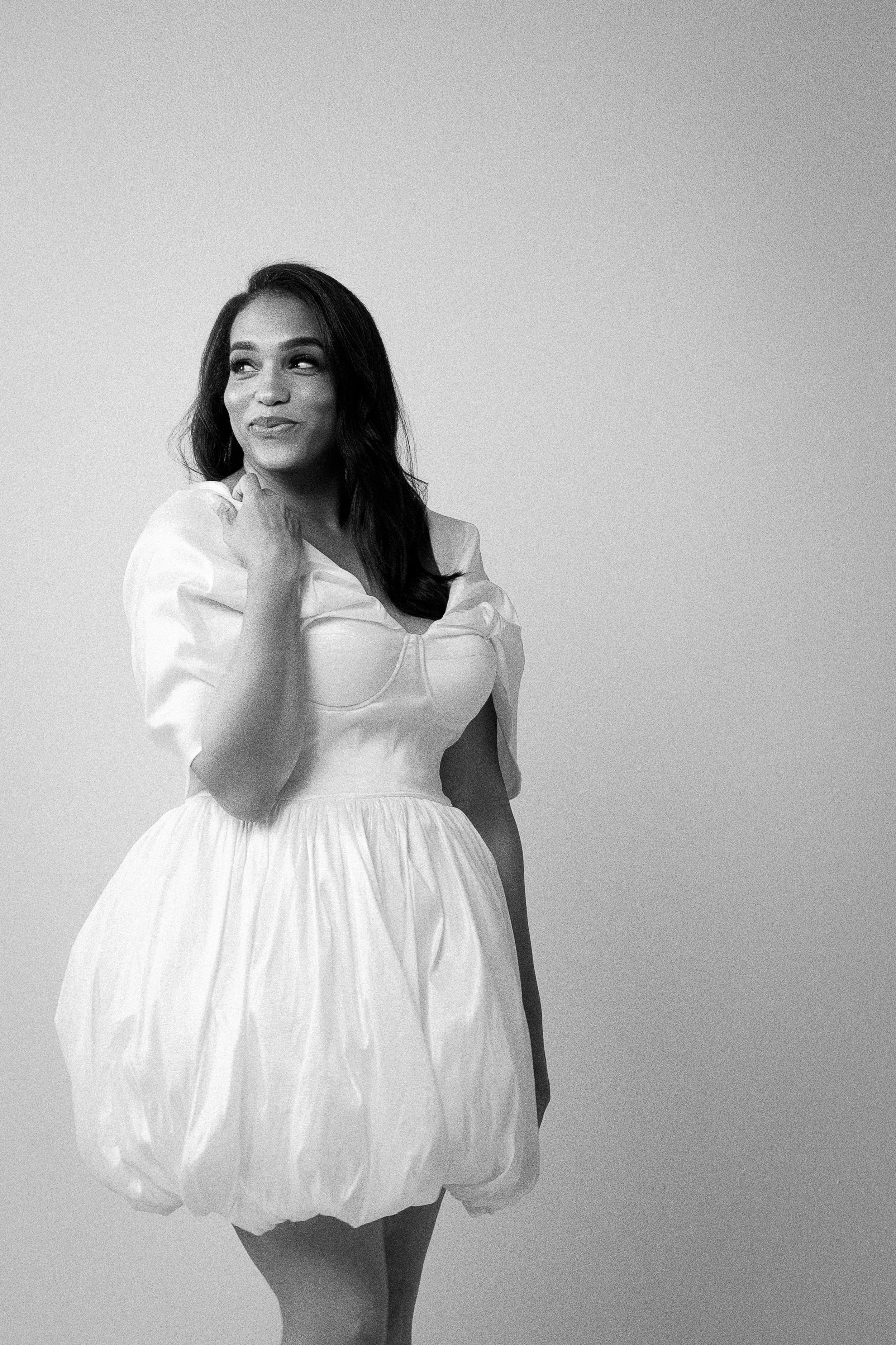 Black and white portrait of a woman with long dark hair wearing a white wedding dress, standing against a plain background.