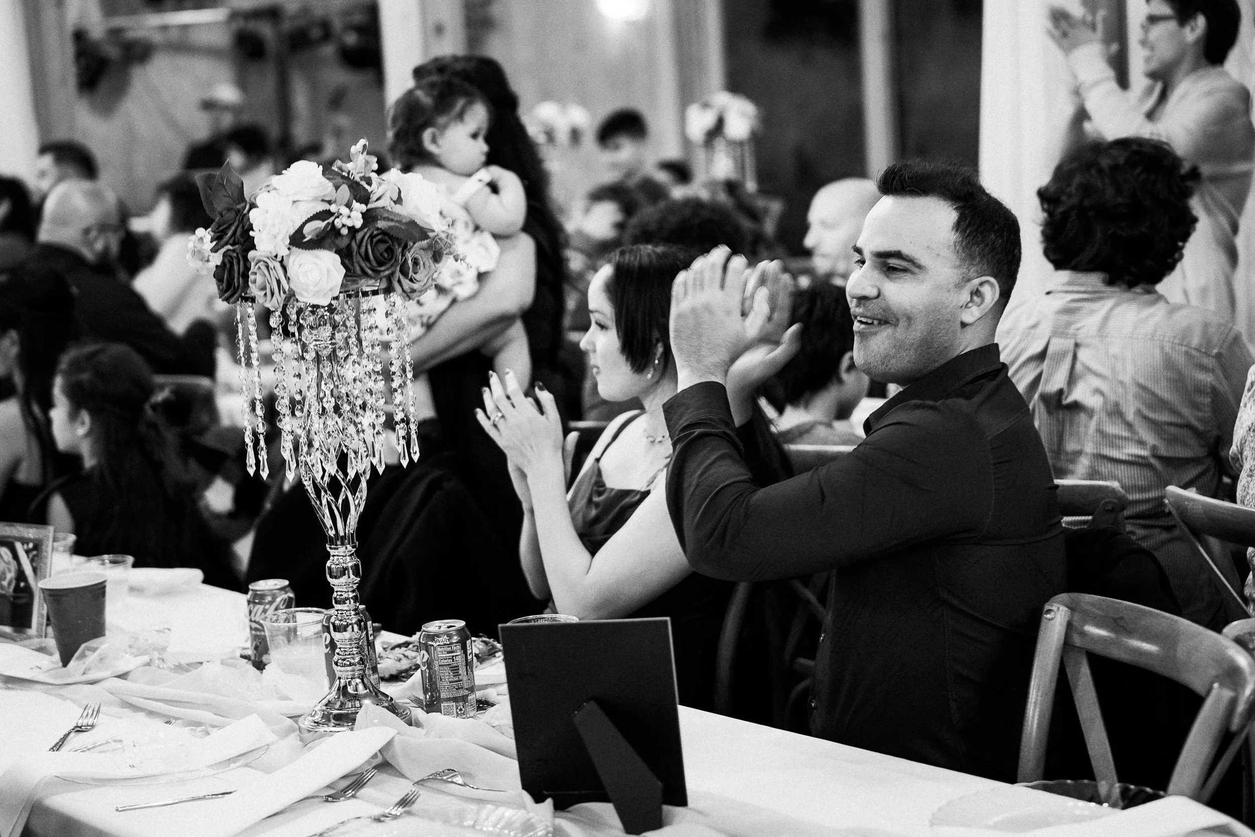 People seated at a wedding banquet table, clapping and smiling, with a decorative vase of flowers and hanging crystal ornaments in the foreground, in a formal event setting.