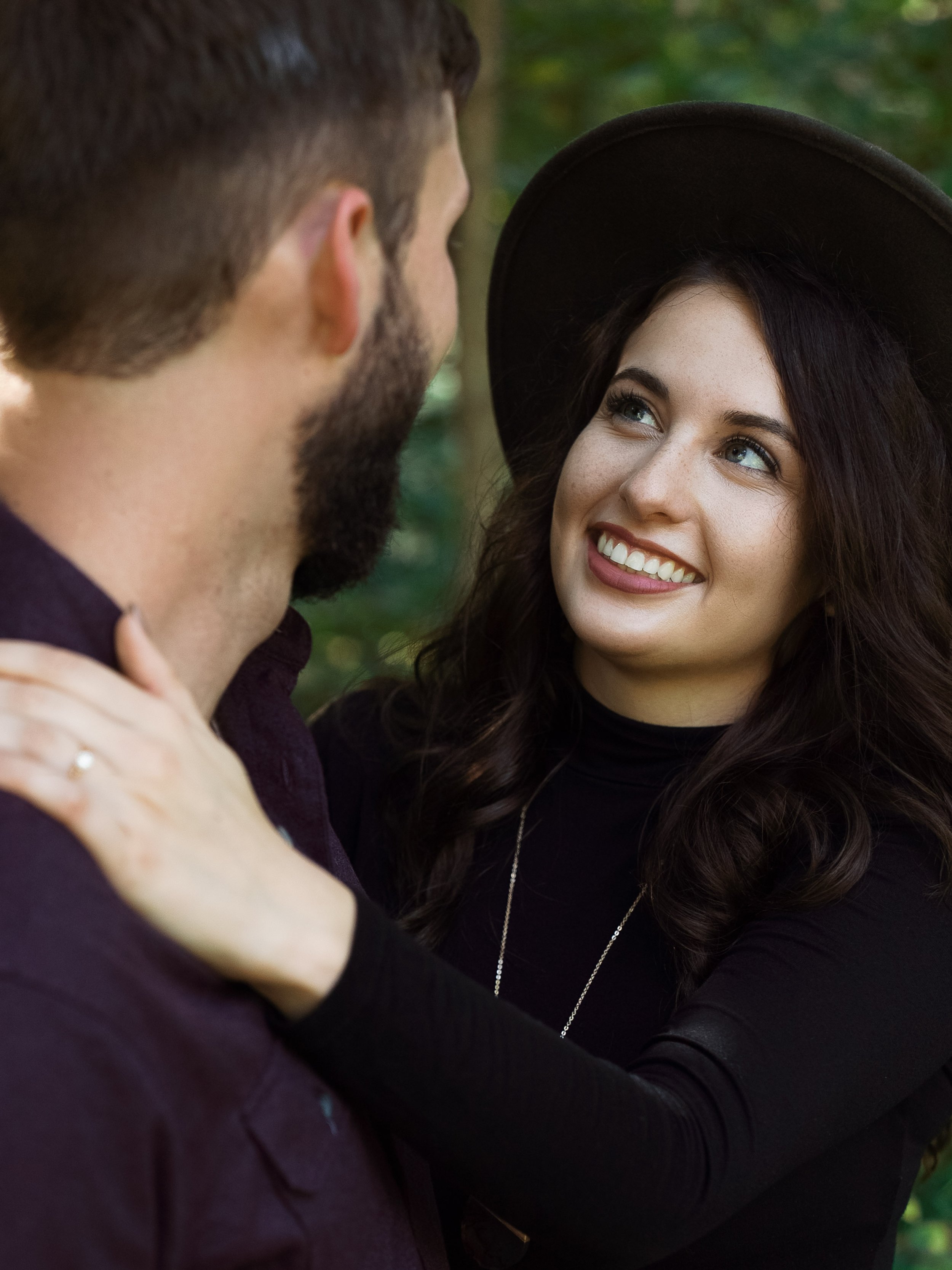 A woman smiling and looking at a man, wearing a black hat and black top, while the man has a beard and is wearing a dark shirt. They are outdoors with greenery in the background.