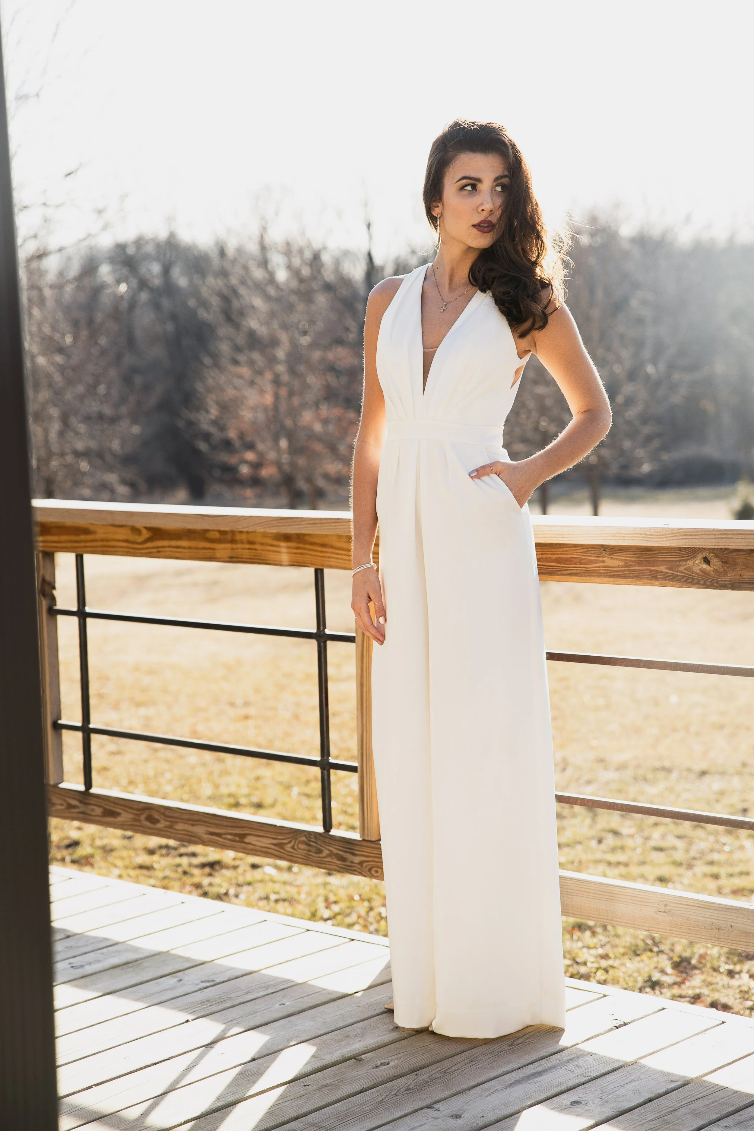 A woman in a white weddig dress standing on a wooden porch with a wooden railing, outdoors with leafless trees in the background and sunlight shining from behind.
