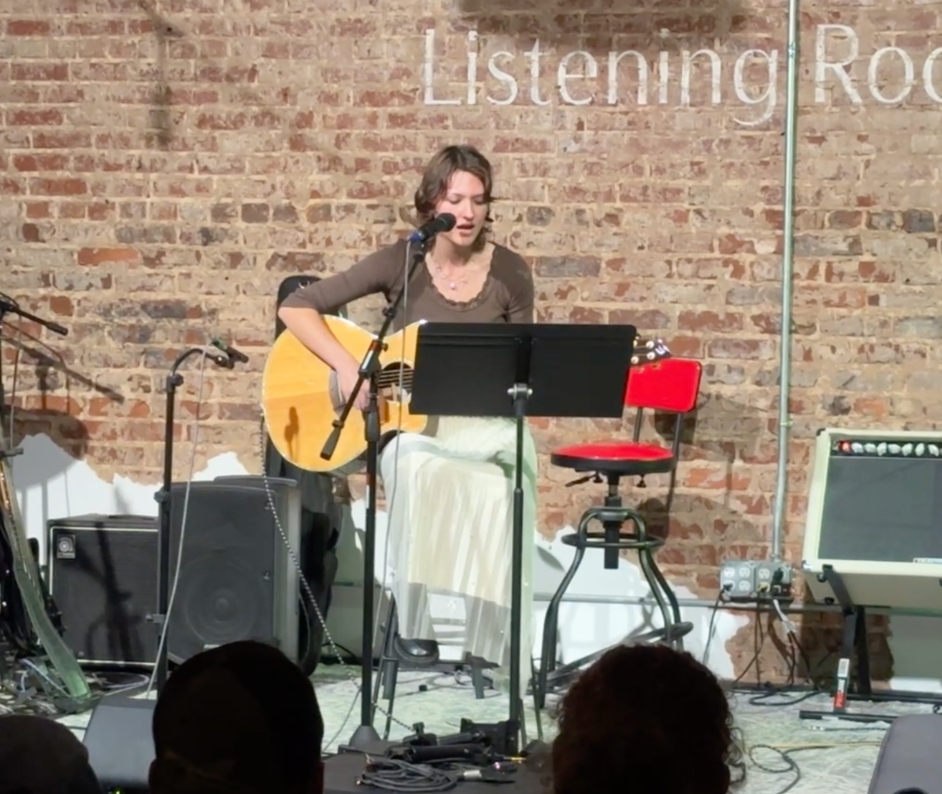 A woman with wavy brown hair performanceing with an acoustic guitar on stage at the Listening Room, with a brick wall background, a music stand, and audio equipment around her.