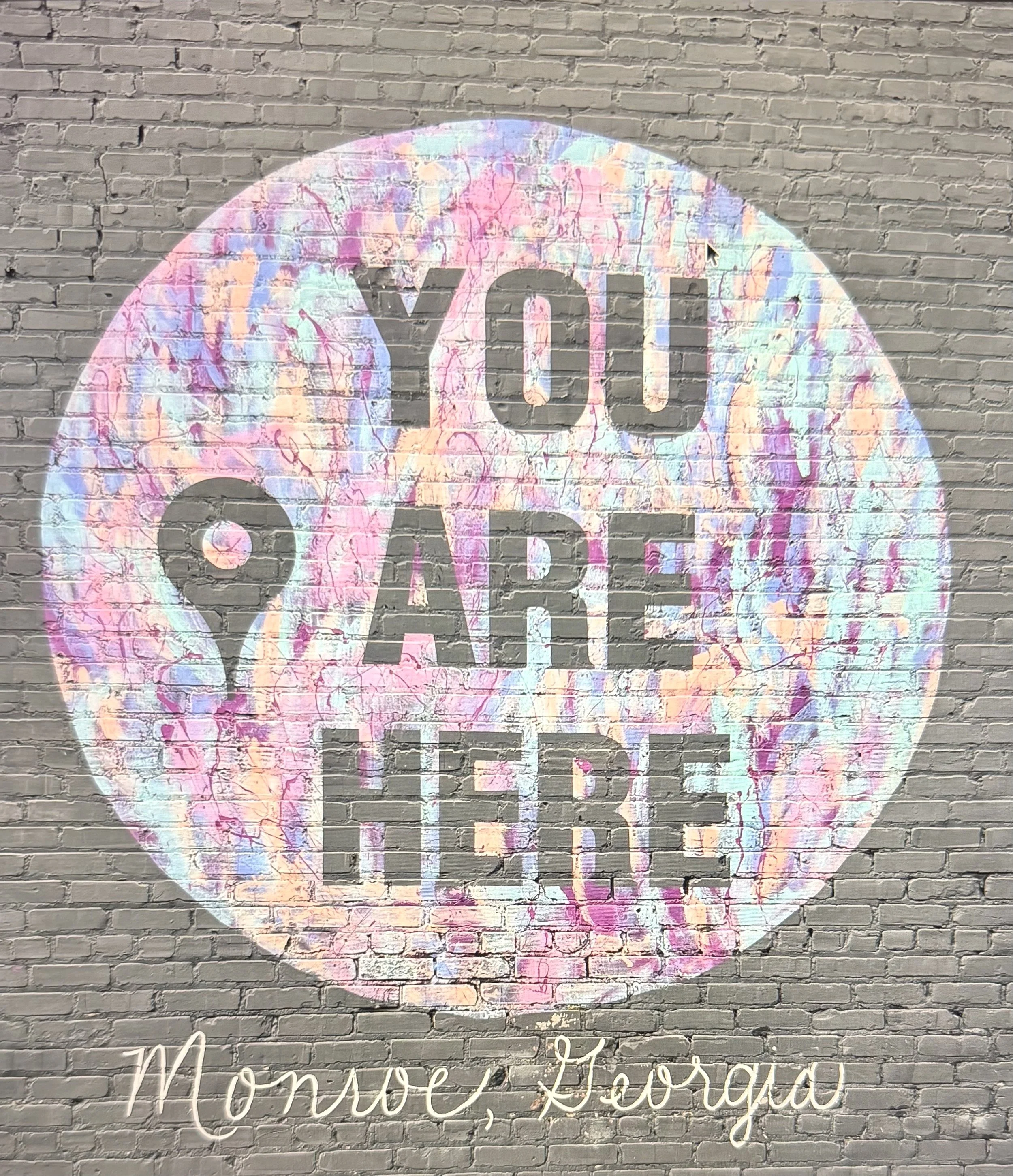 A mural on a brick wall with colorful pastel swirls and the message 'You Are Here' in bold, gray letters, and the location 'Monroe, Georgia' written in white cursive at the bottom.
