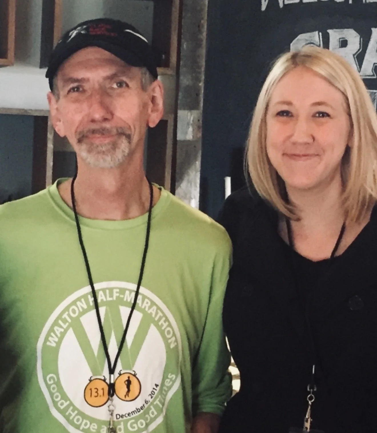 A man and woman standing indoors, smiling at the camera. The man is wearing a bright green marathon shirt, a black cap, and a medal around his neck. The woman has blonde hair, wears a black top, and has a lanyard with keys around her neck.