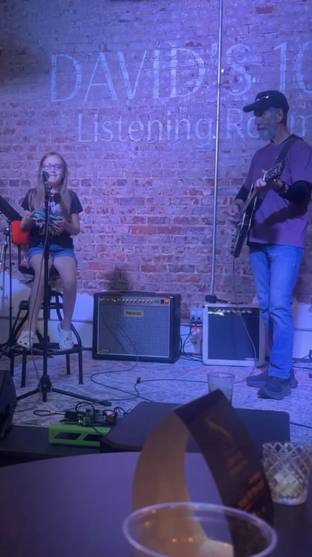 A girl sitting on a chair singing into a microphone and a man playing an acoustic guitar on stage against a brick wall with lighting that says 'Davis Listening Room'.
