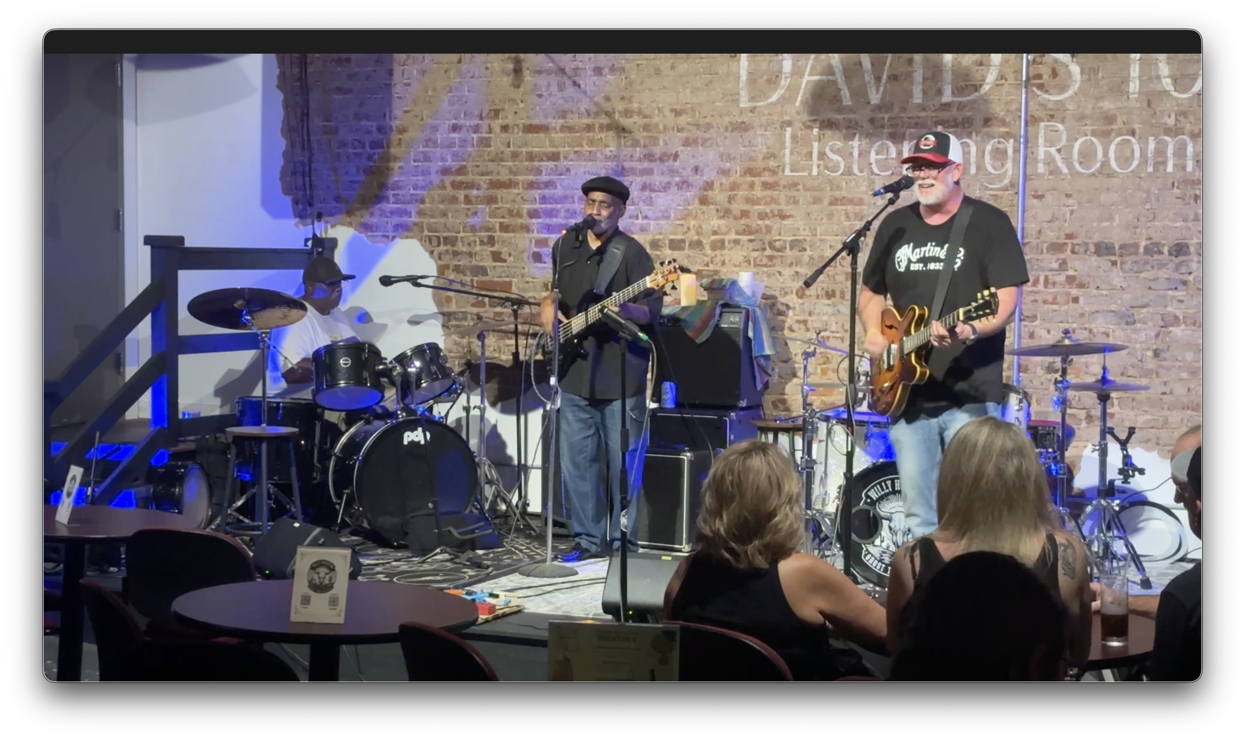 Band performing on stage at Davidson Listening Room, with three musicians playing guitar, bass, and drums, against a brick wall, with audience seated in front.
