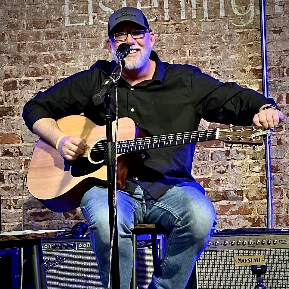 Rod Avera playing guitar with microphone seated on stool in front of a brick wall and amps at David's 105 Listening Room.