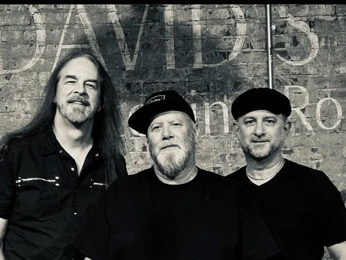 Three men standing in front of a brick wall with partly visible text.