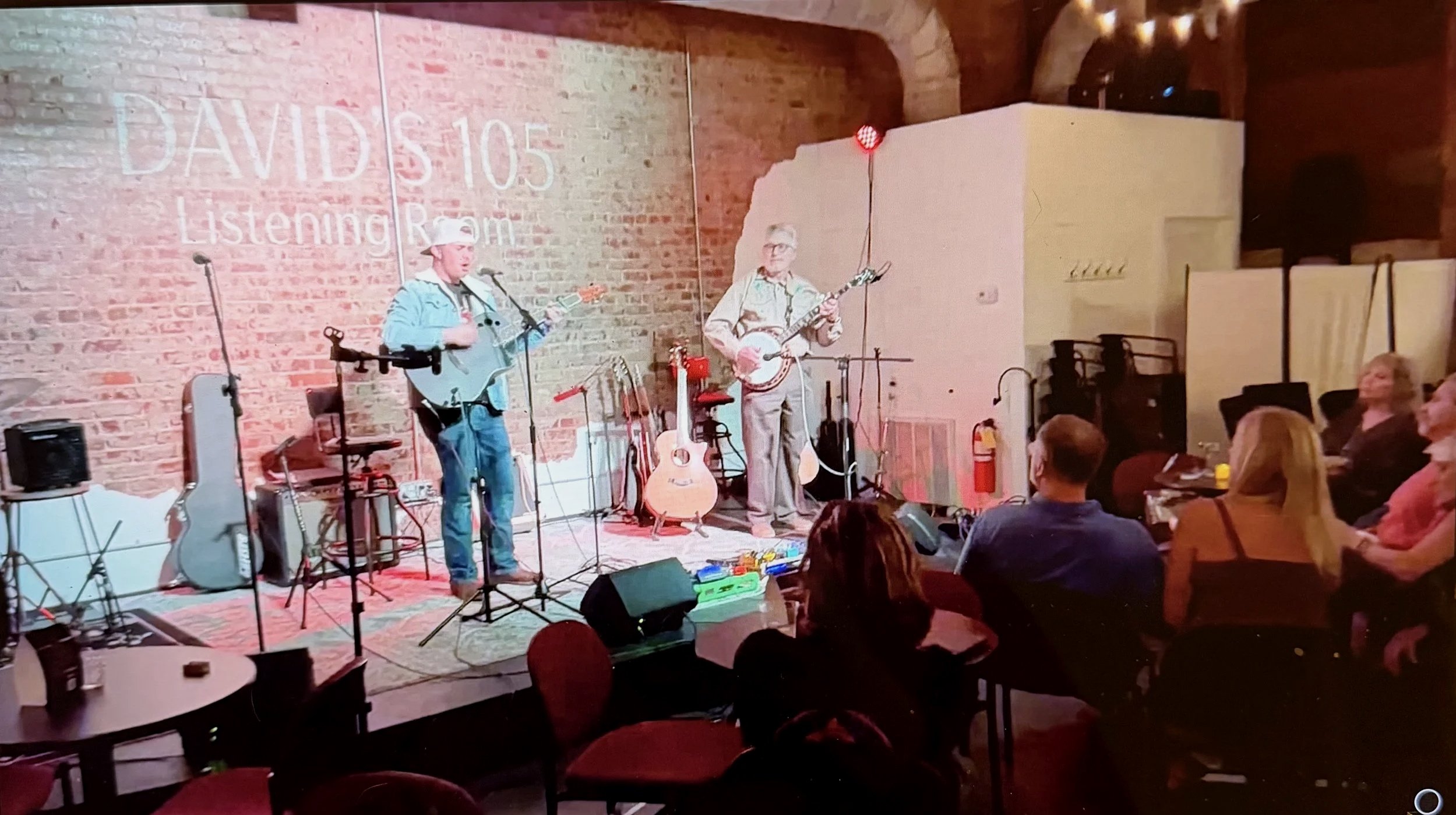 Two musicians perform on stage in a brick-walled venue, one playing guitar and the other playing a banjo, with audience members seated and watching.