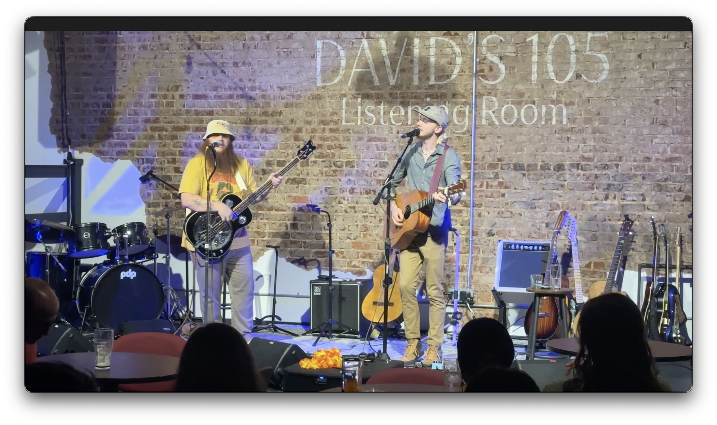 Two musicians perform on stage at Davis 105 Listening Room, one playing guitar and singing, the other playing a bass guitar and singing, with guitars and musical equipment surrounding them, an audience watching in the foreground.