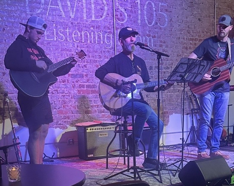 Three men performing acoustic music on stage with microphones and guitars, against a brick wall background.