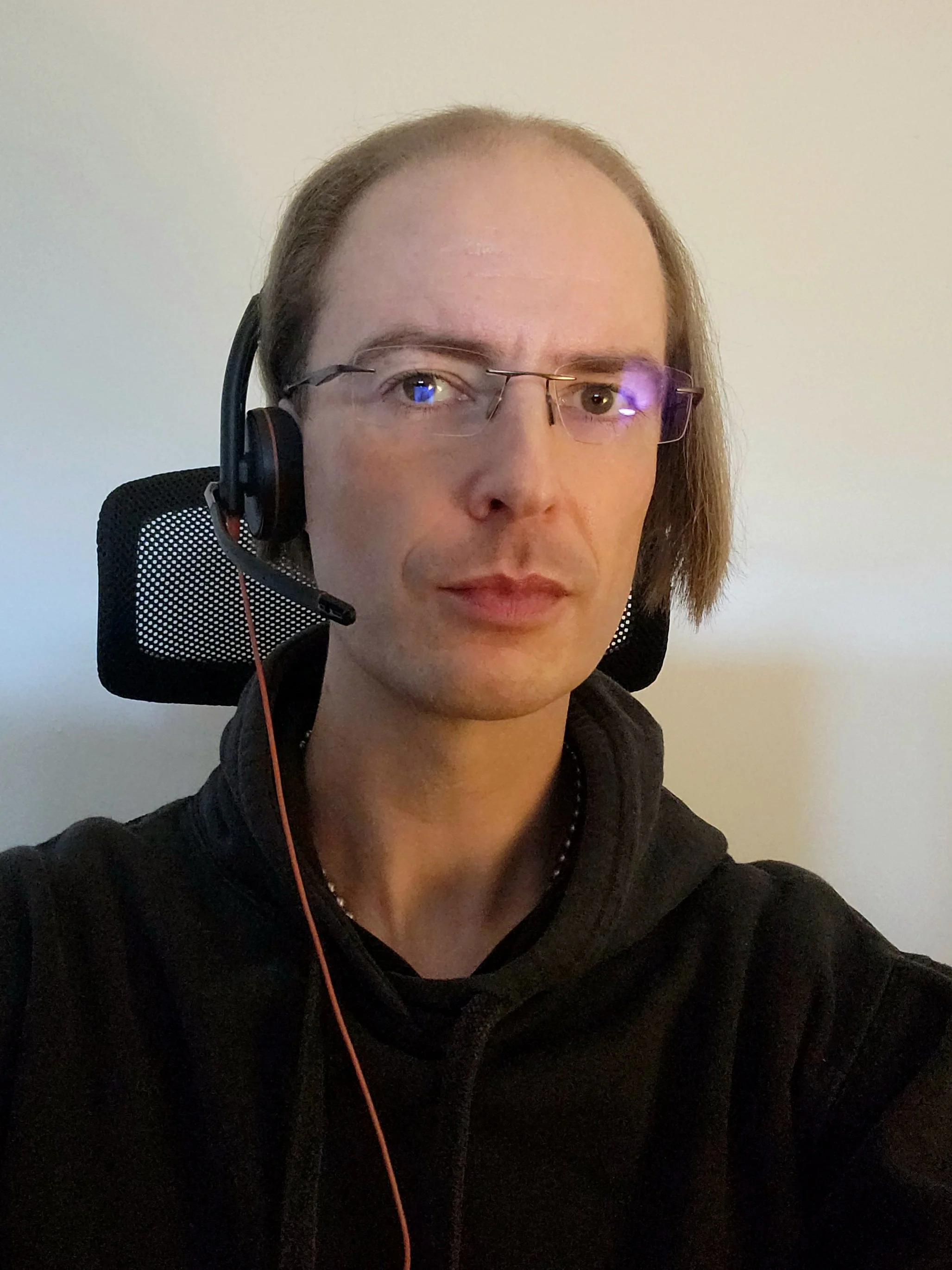 A person with glasses wearing a black hoodie sitting in front of a plain wall, using a headset with a microphone.