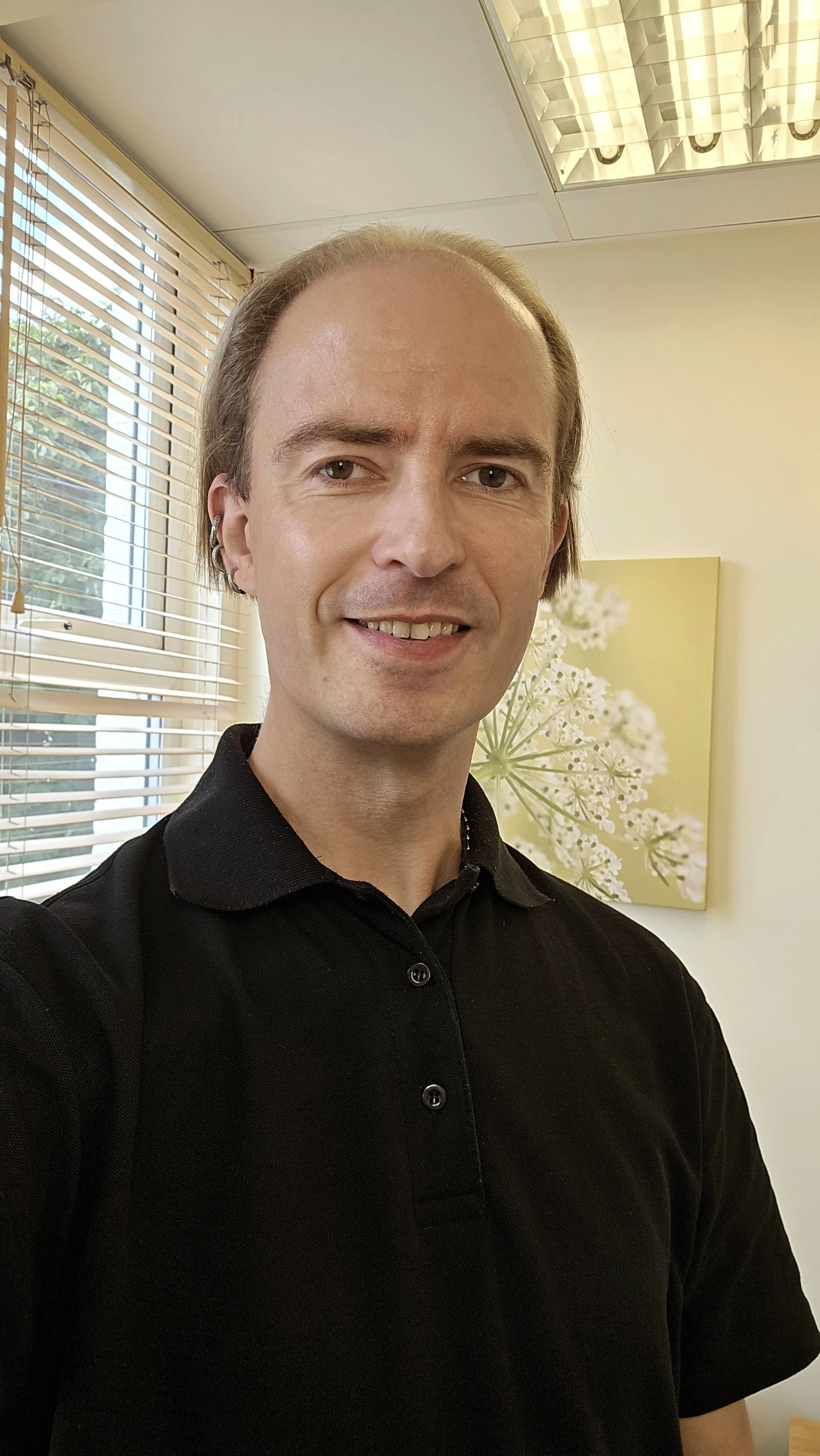 A man with light brown hair, styled back, wearing a black polo shirt, standing indoors near a window with blinds. There is a floral painting or photograph on the wall behind him.