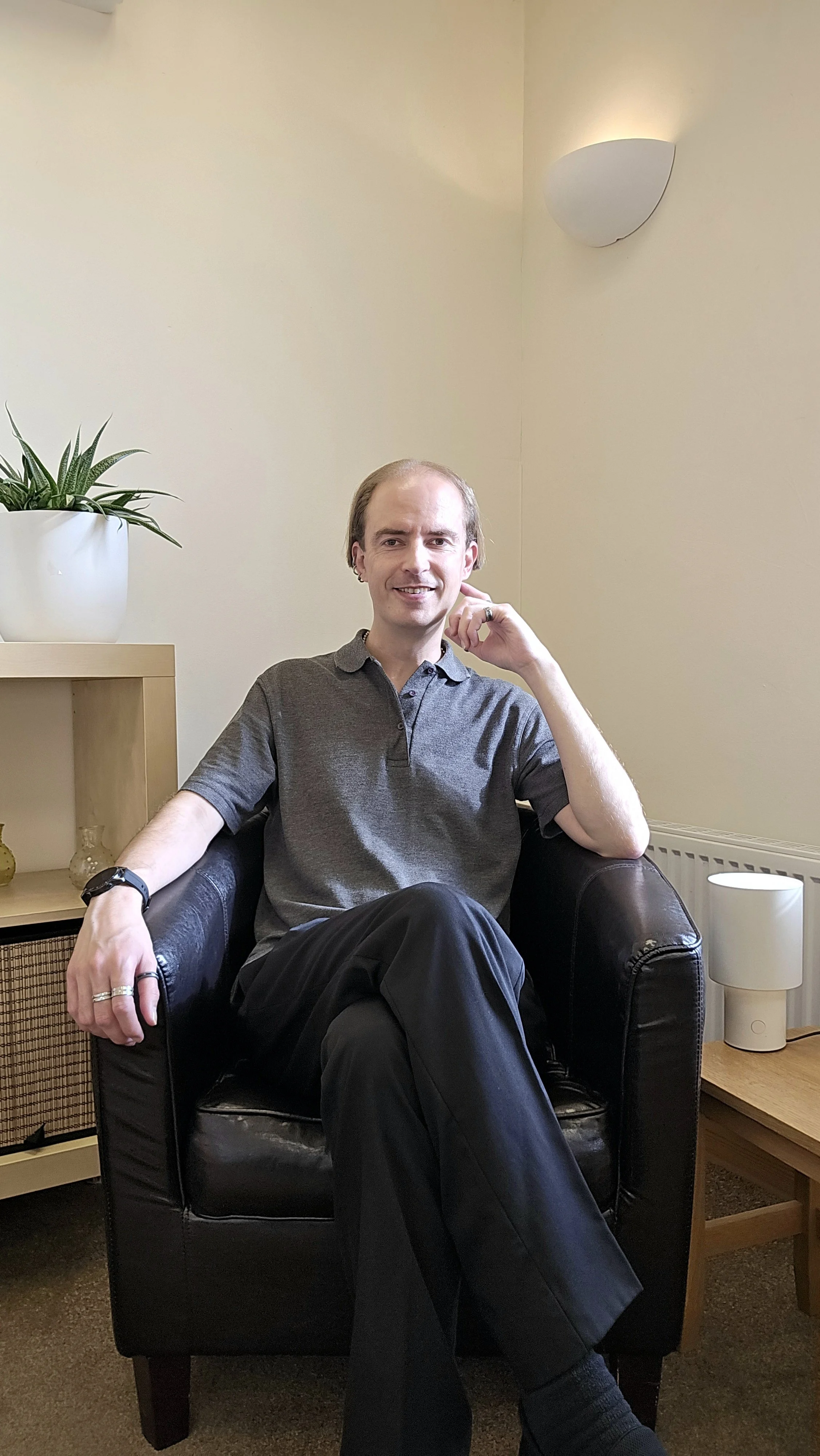 A man with light skin, light hair, and earrings sitting in a black leather armchair in a room with beige walls. He is wearing a gray polo shirt, black pants, and has a watch and rings on his left hand. There is a white plant pot with a green plant on a wooden shelf to his left, and a small white lamp on a wooden side table to his right. A wall-mounted light fixture is visible on the wall.