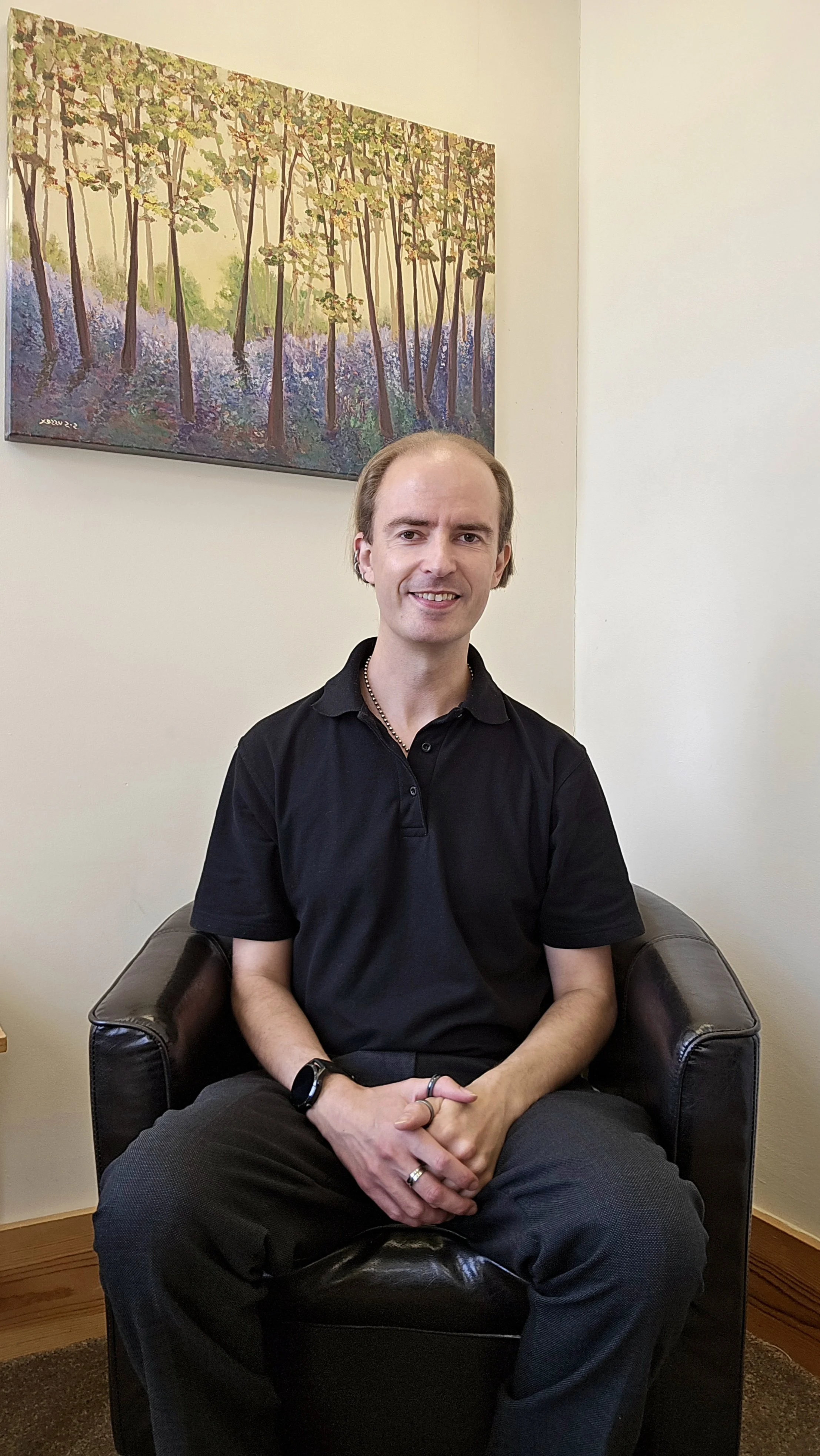 A man with light skin, wearing a black polo shirt, black pants, and a watch, sitting on a dark leather armchair in a room with beige walls and hardwood flooring. Behind him, a colorful painting of a forest scene with trees and purple flowers hangs on the wall.